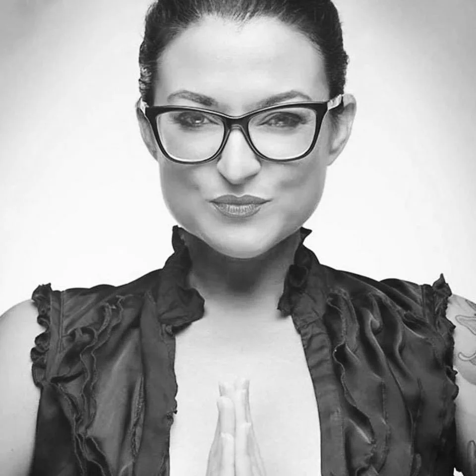 A black and white photo of a woman with short dark hair wearing glasses and a sleeveless ruffled blouse, smiling at the camera.