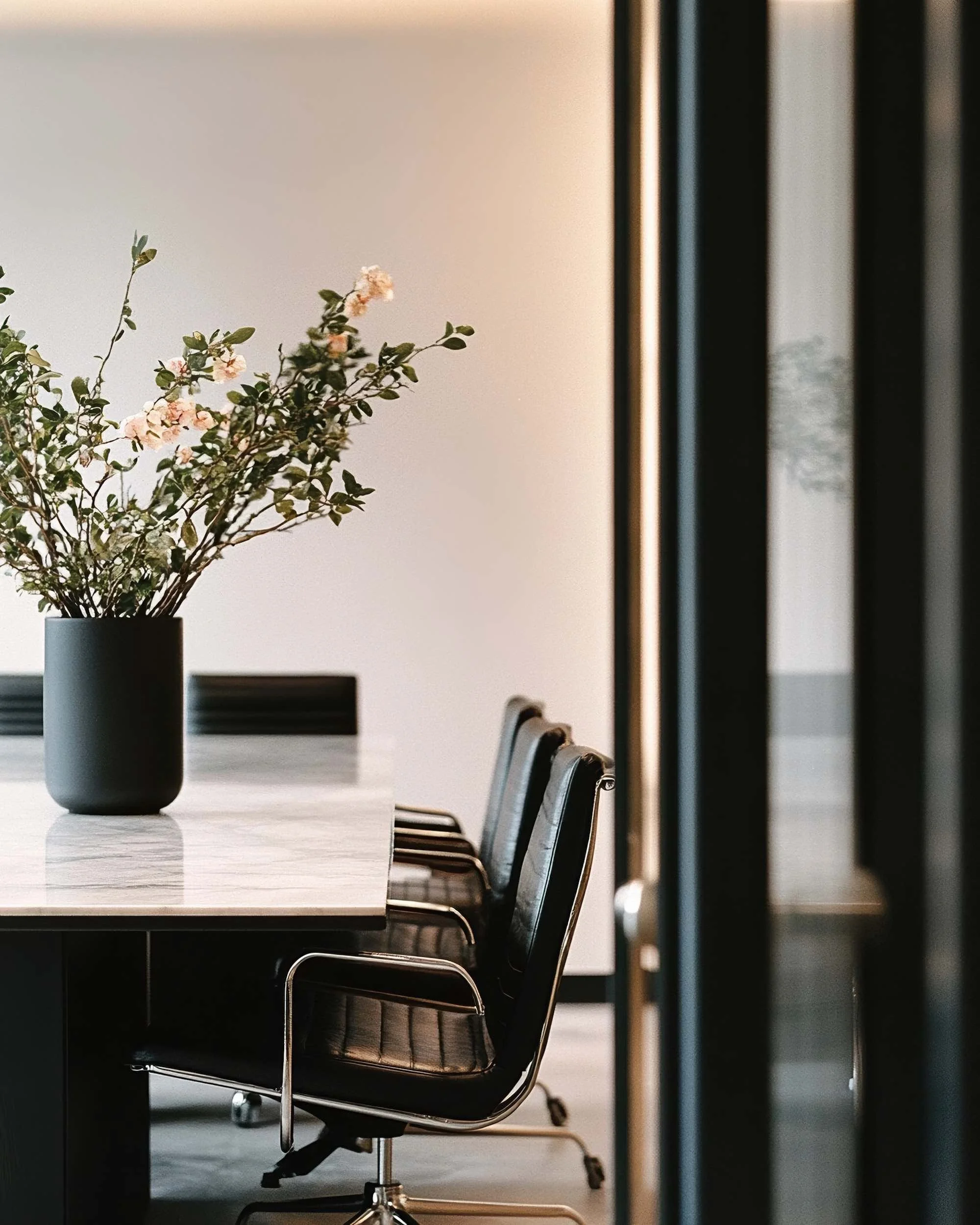 An office conference room with a long wooden table, black leather chairs, and a white vase with pink and white flowers on the table. Sunlight filters through the windows, creating a bright and modern workspace.