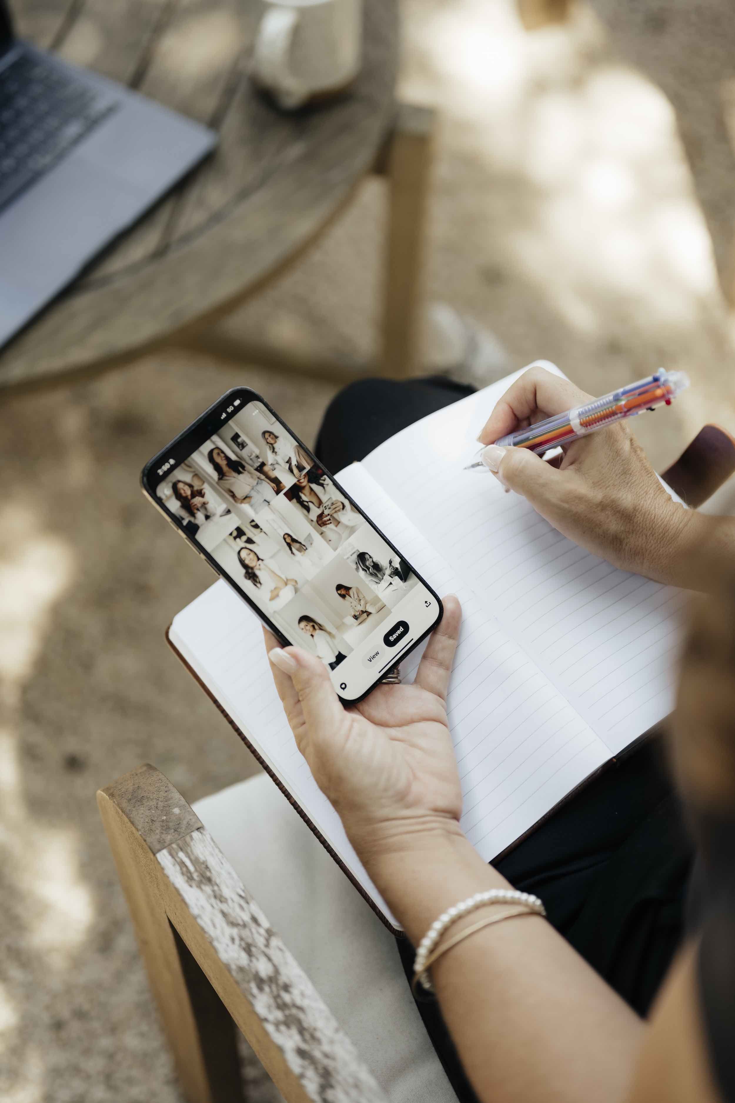 A person sitting at a wooden outdoor table, holding a smartphone displaying a collage of images of women, with a pen and notebook in their other hand, wearing a pearl bracelet.