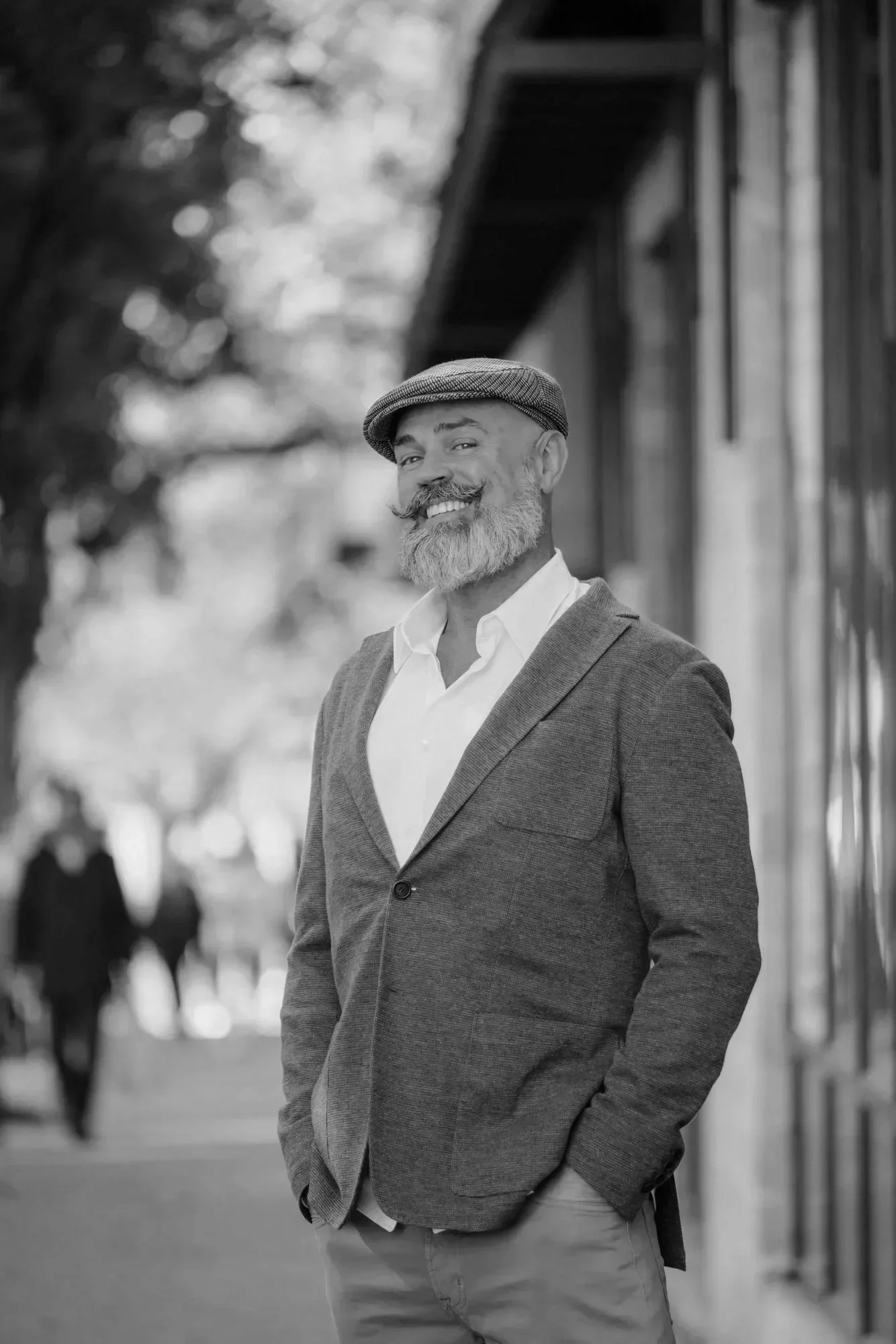 Man smiling outdoors in a suit jacket and flat cap, standing near a building with blurred figures in the background.
