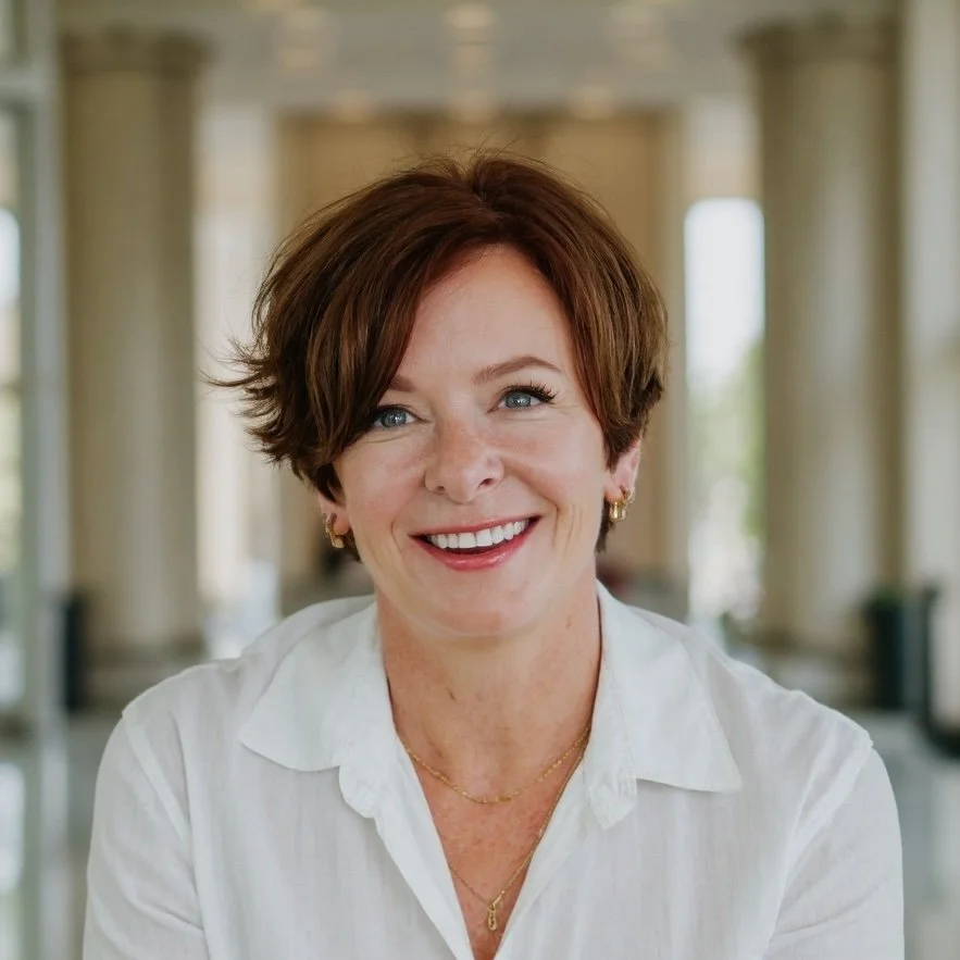 A woman with short brown hair, wearing a white shirt, smiling, indoors with columns in the background.