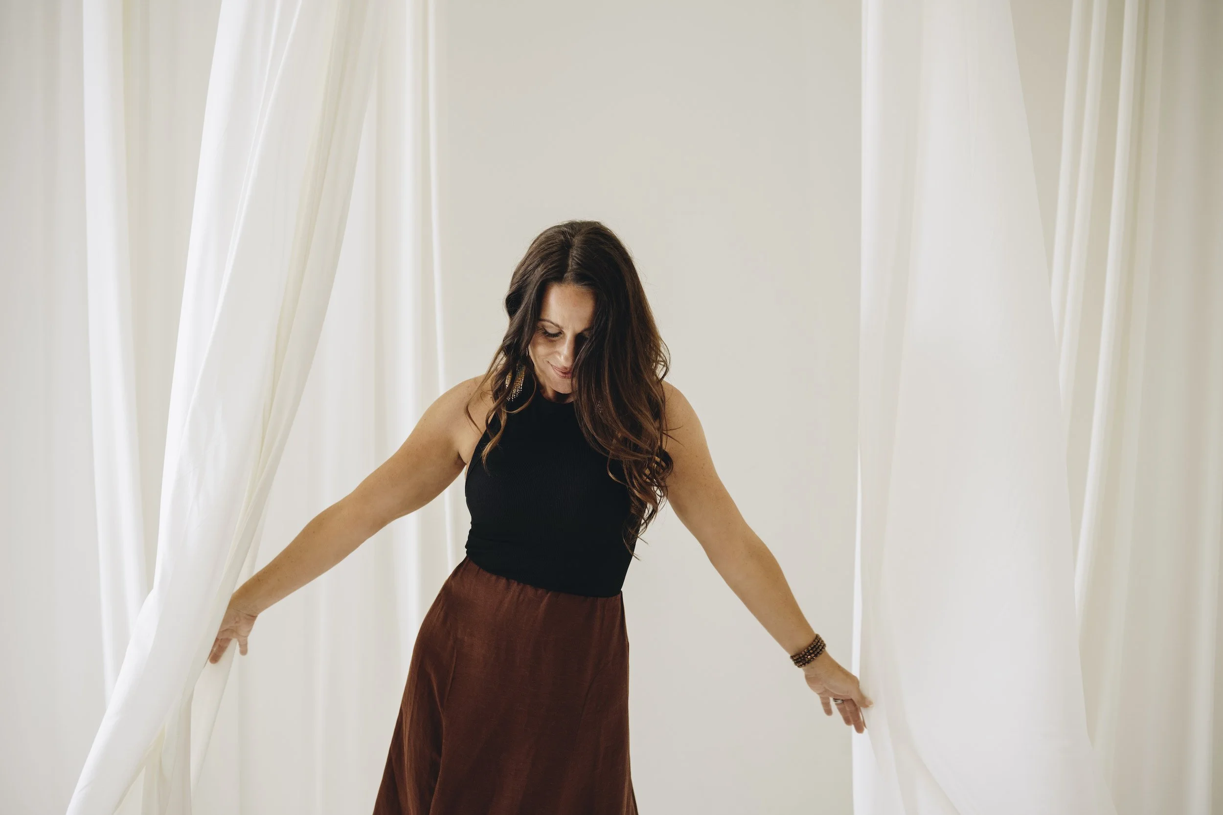 Woman with long wavy hair smiling, wearing a black sleeveless top and a brown skirt, standing between white curtains, leaning slightly forward with arms extended.