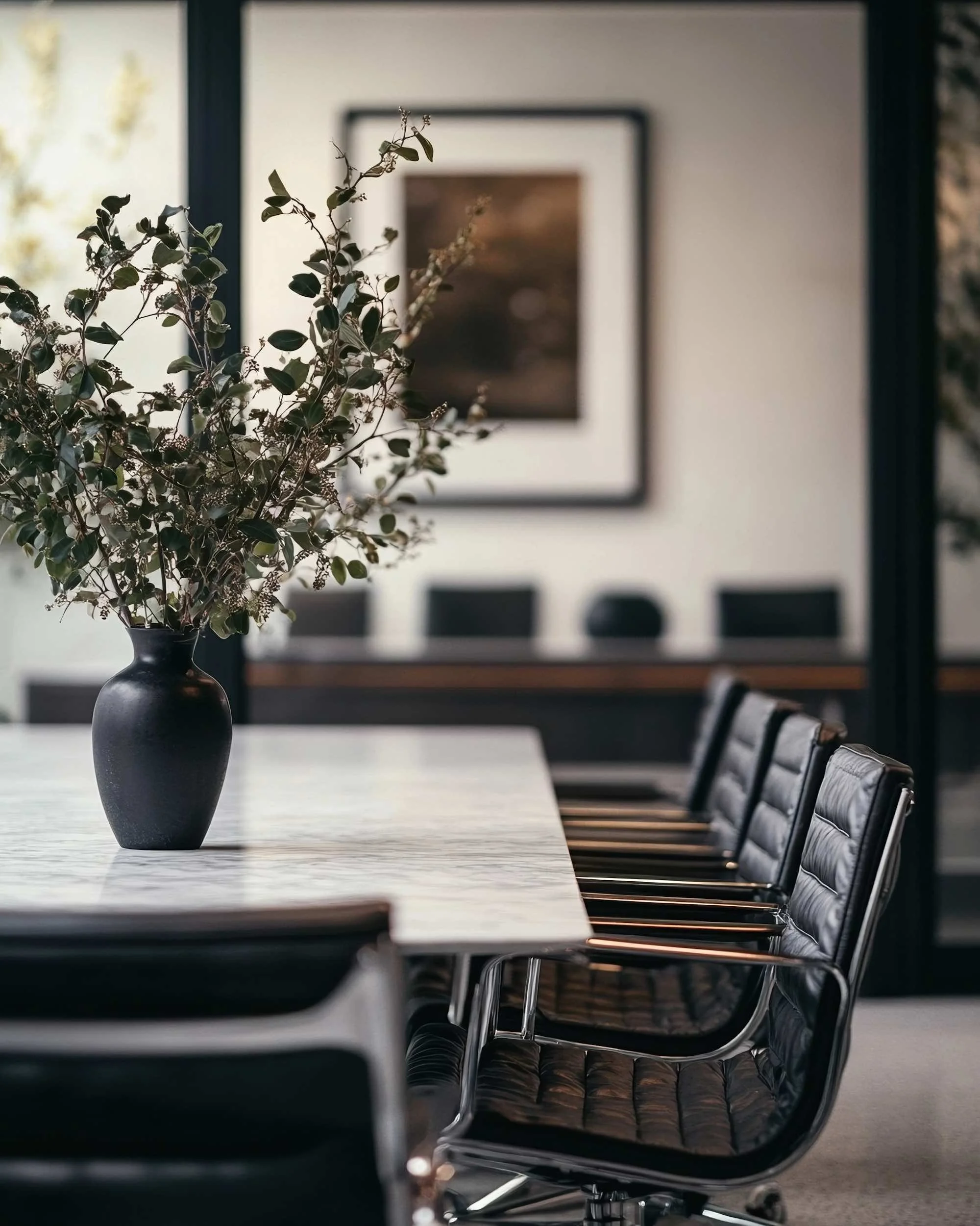 A black vase with dried green leaves and small flowers sits on a white marble table in a modern room with black chairs and framed artwork on the wall in the background.