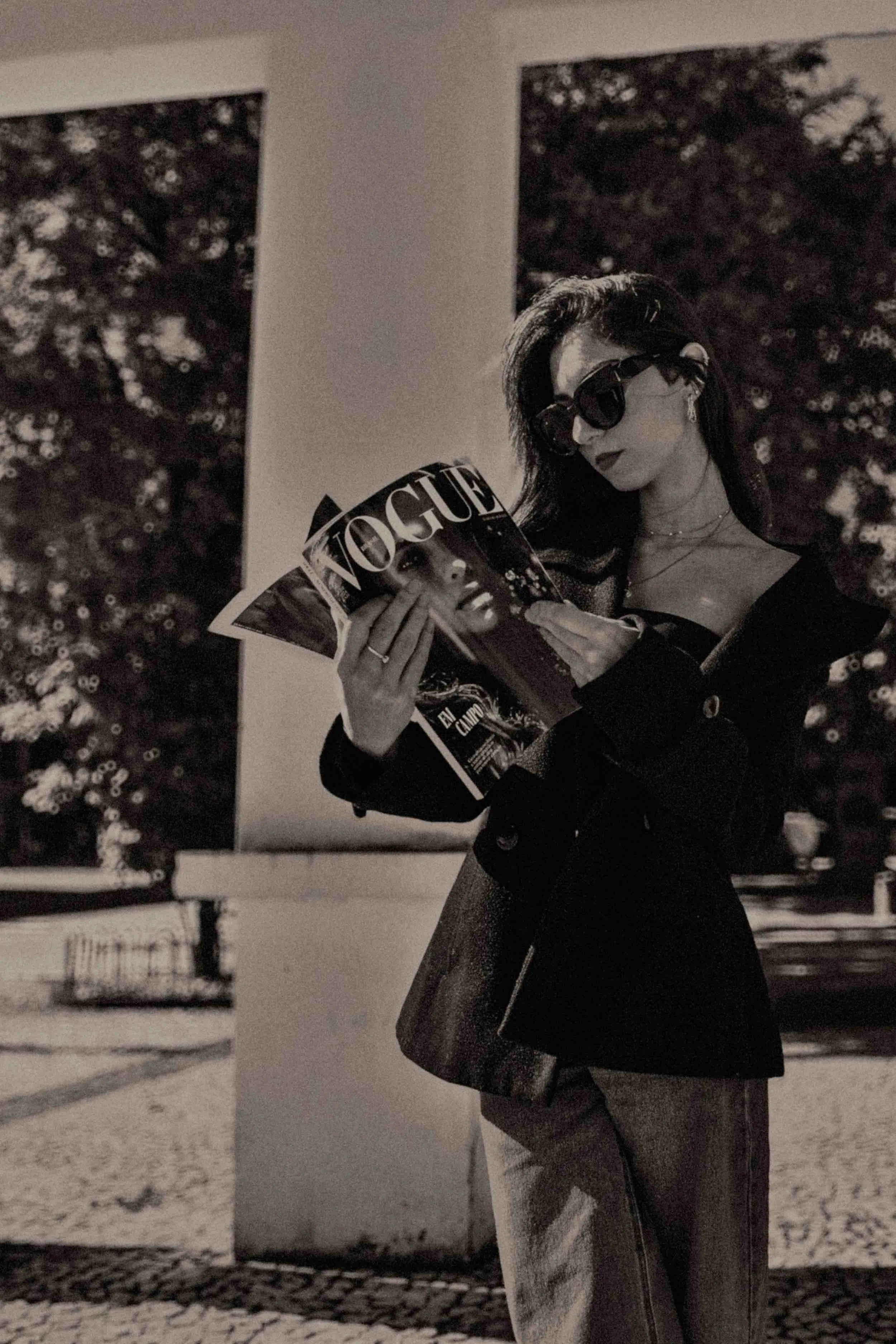 A woman with dark hair wearing sunglasses reading a Vogue magazine outdoors in a park with trees and a bench in the background.