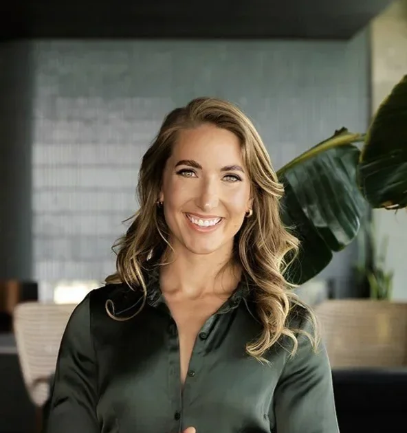 A smiling woman with wavy hair wearing a dark green button-up shirt, standing indoors with a large green plant behind her and modern decor.