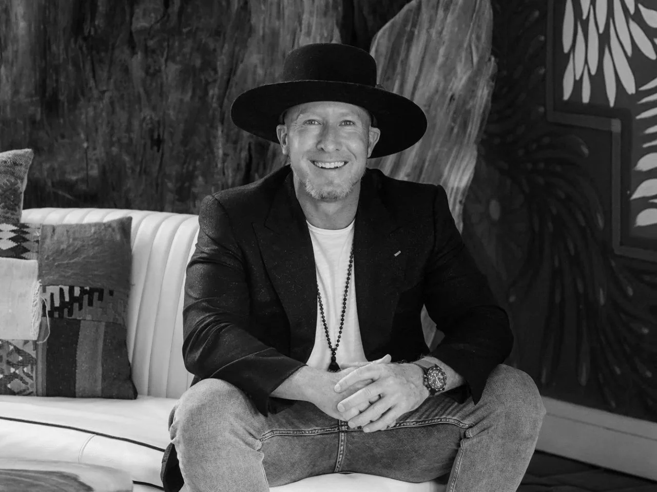 Man sitting on a sofa, smiling, wearing a wide-brimmed hat, blazer, and jeans, with a decorative background and pillows.