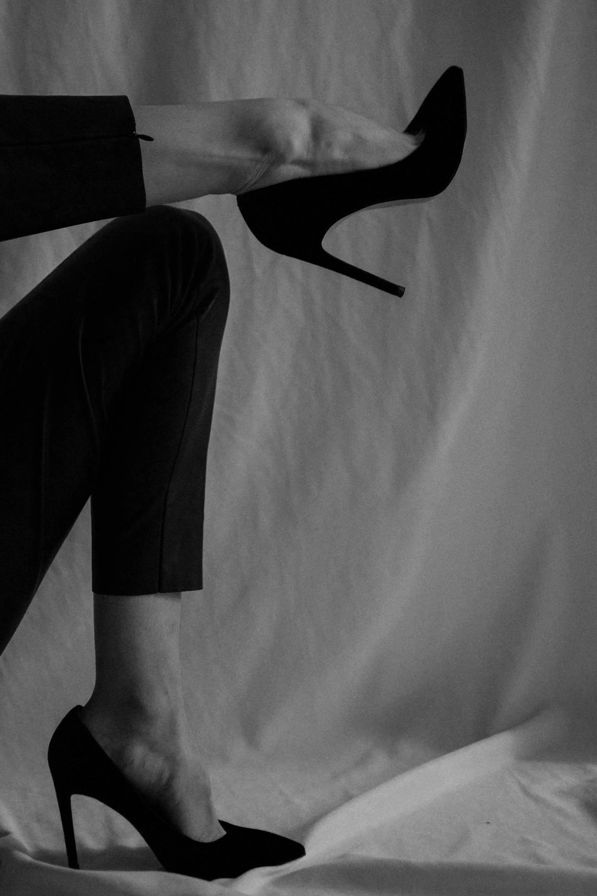 Black and white photo of a woman in high heels, sitting with one leg crossed over the other, with a plain backdrop.