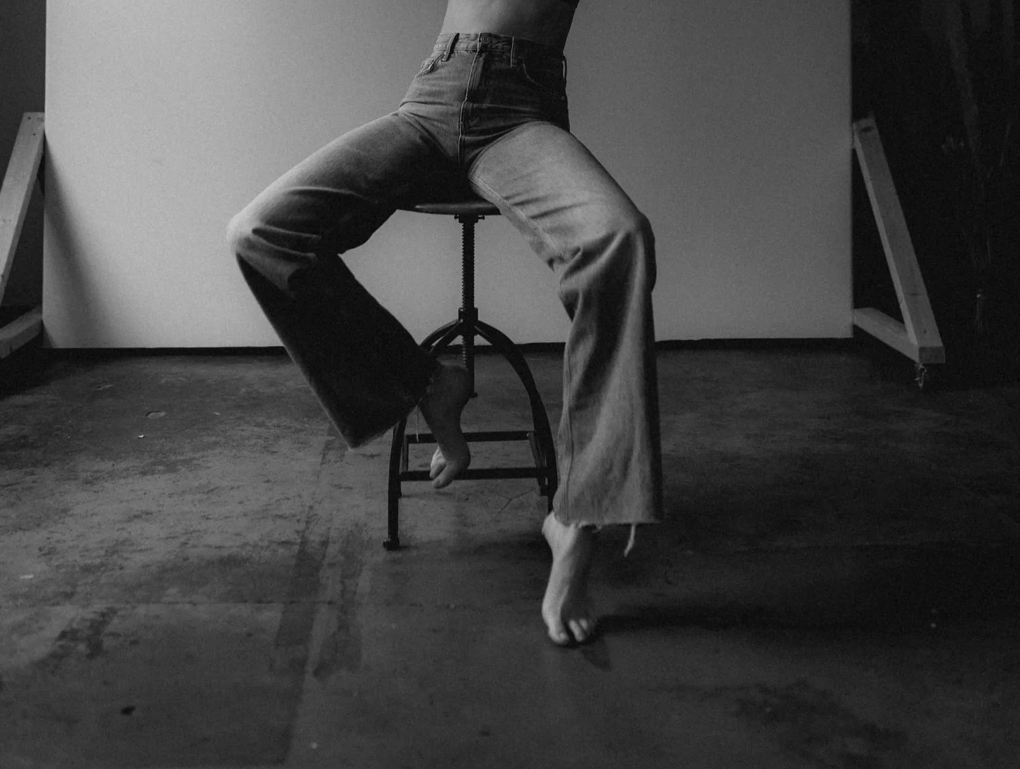 Black and white photograph of a person sitting on a stool, with legs apart, wearing vintage jeans. The person's face and upper body are not visible. The setting appears to be an art studio or photo shoot space, with a plain background and some wooden structure on the sides.