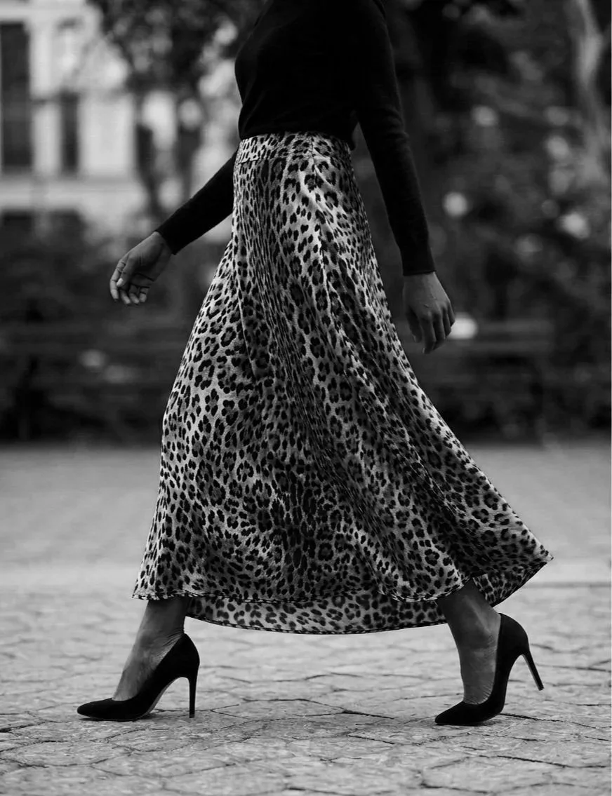 Black and white image of a woman walking, wearing a leopard print skirt, black top, and high heels, with background trees and blurred buildings.