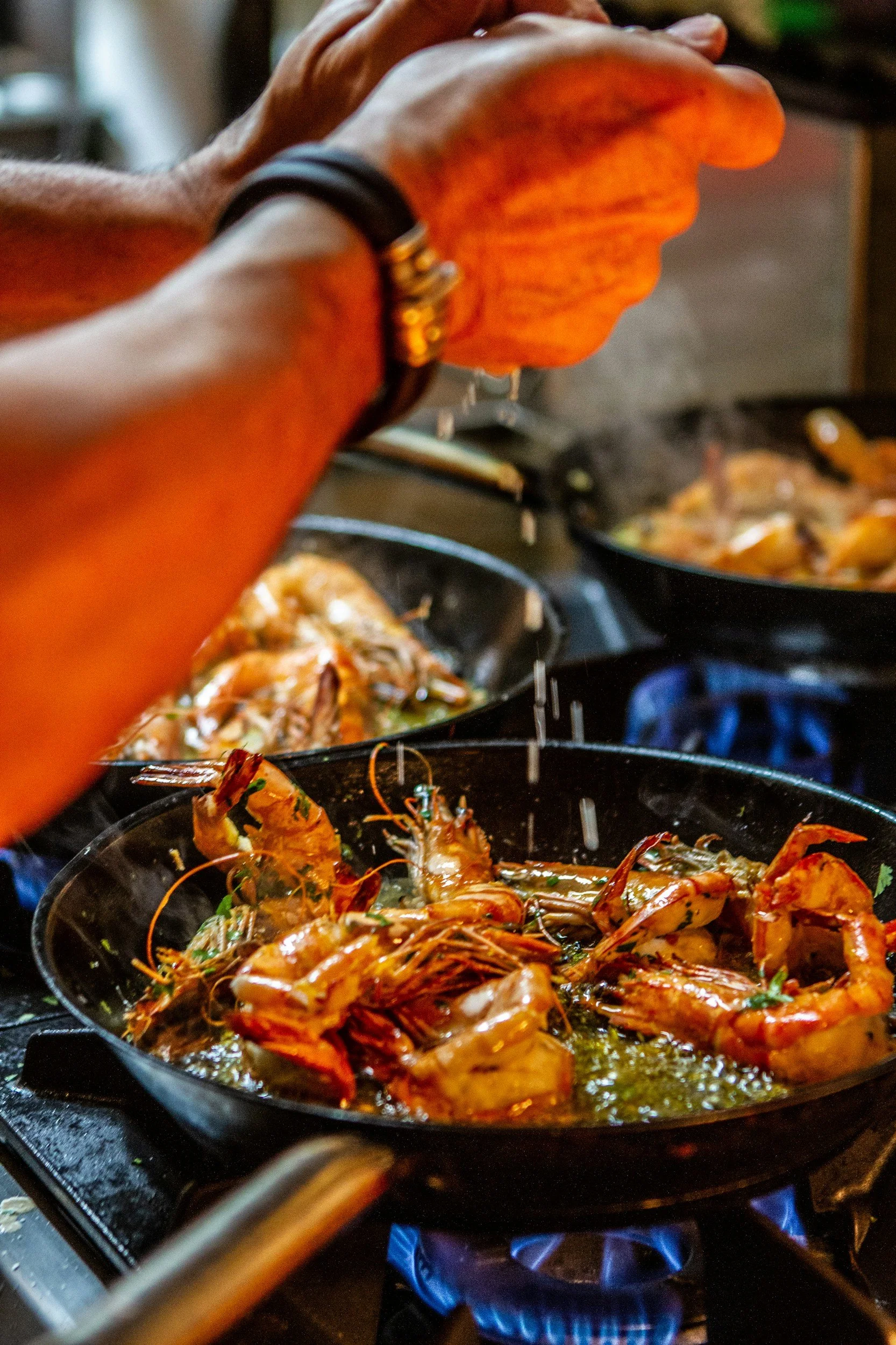 Cooking shrimp in a black skillet on a stovetop with blue flames, with another pan of food in the background.