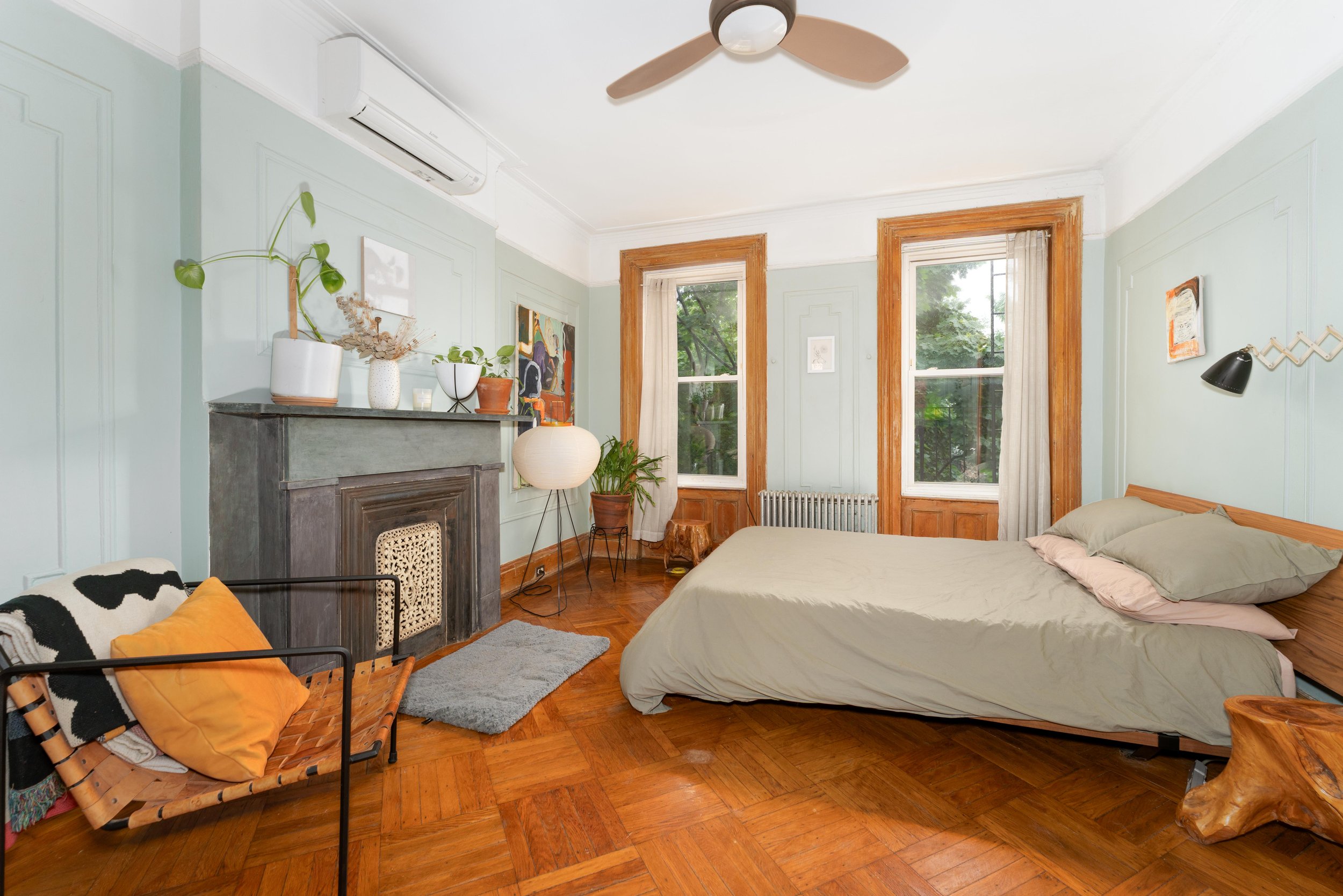 A bedroom with two large windows, light green walls, and wooden trim. There is a bed with beige bedding, a side table with plants and artwork on the wall, a vintage-style fireplace, and a woven lounge chair with orange cushions. A ceiling fan is attached to the ceiling.