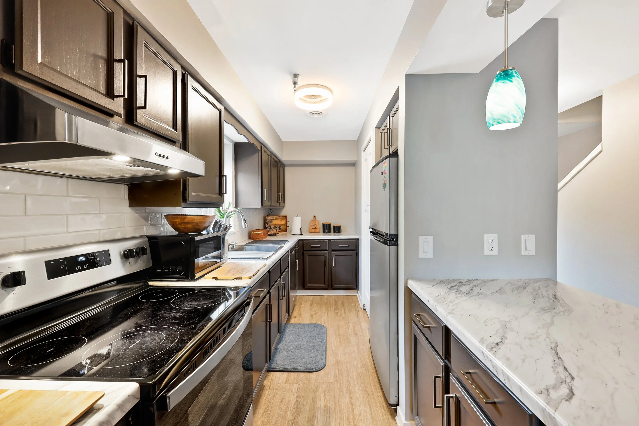 A modern kitchen with dark brown cabinets, stainless steel appliances, a marble countertop, and a hanging blue-green pendant light.
