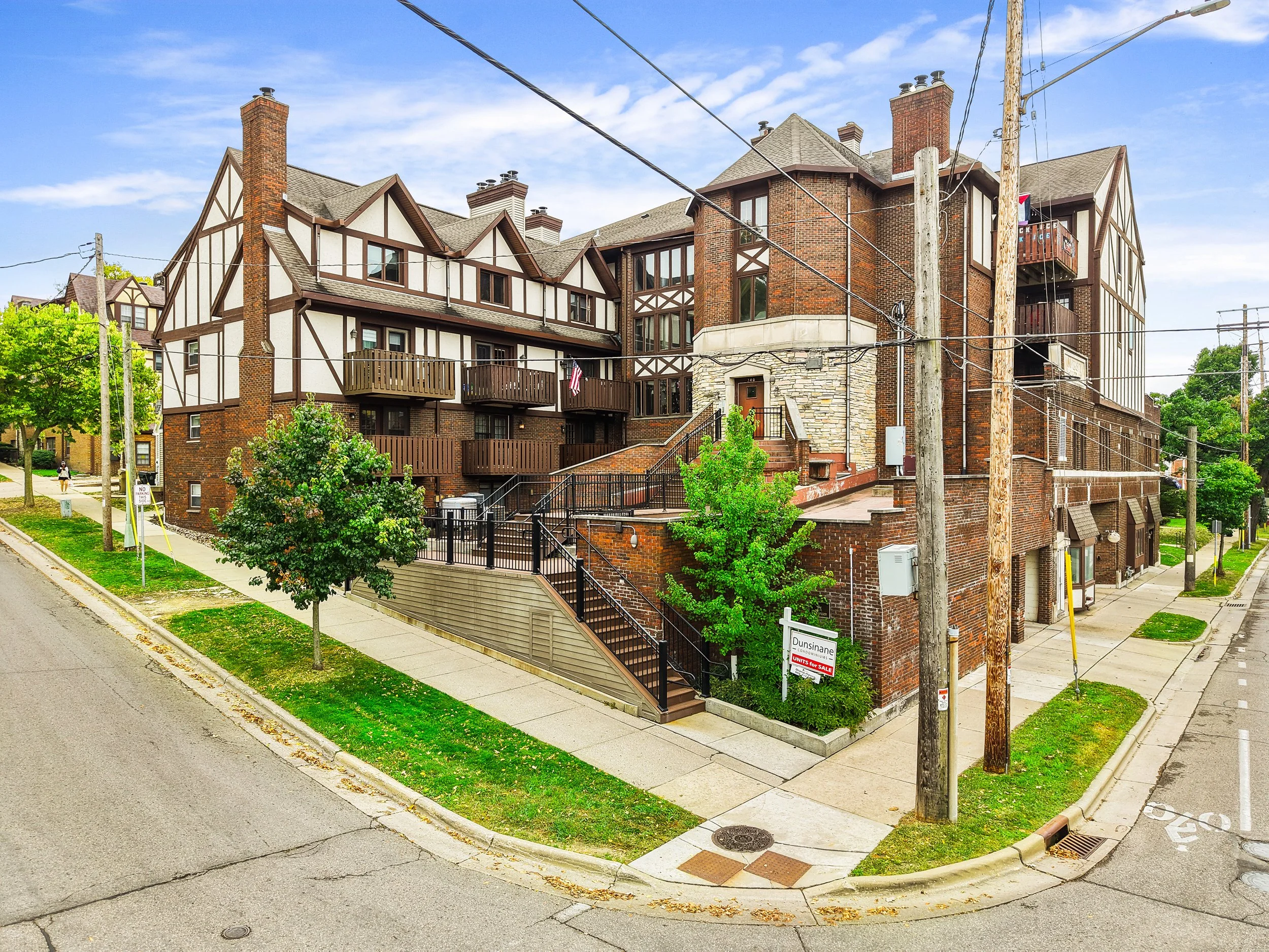 A multi-story brick and half-timbered apartment building on a street corner with a sidewalk, green trees, utility poles, and power lines in front. There is a sign indicating units for sale.
