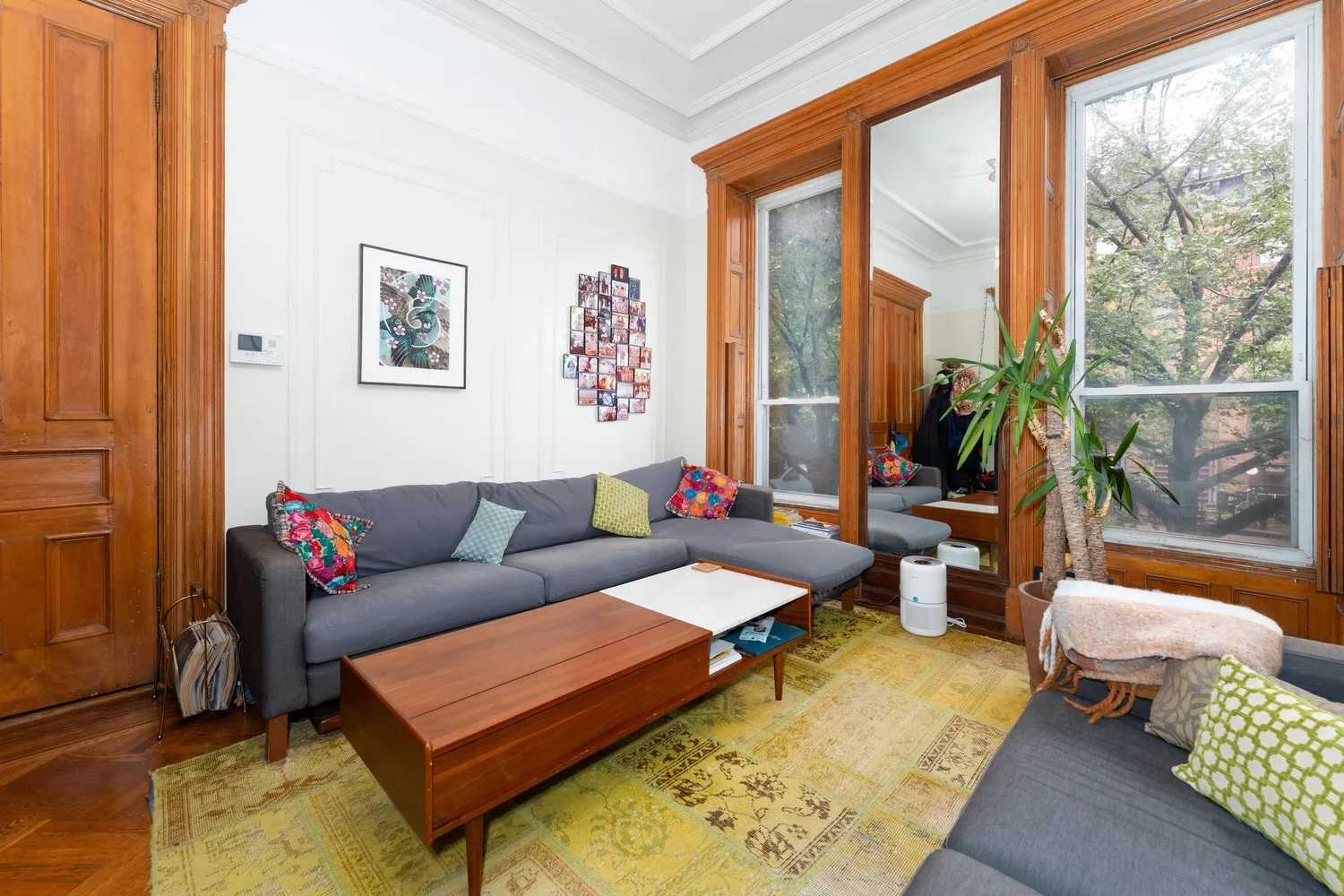 Living room with white walls, wooden trim, large windows, gray sectional sofa with colorful pillows, wooden coffee table, patterned rug, potted plant, artwork on wall, and a small white air purifier.