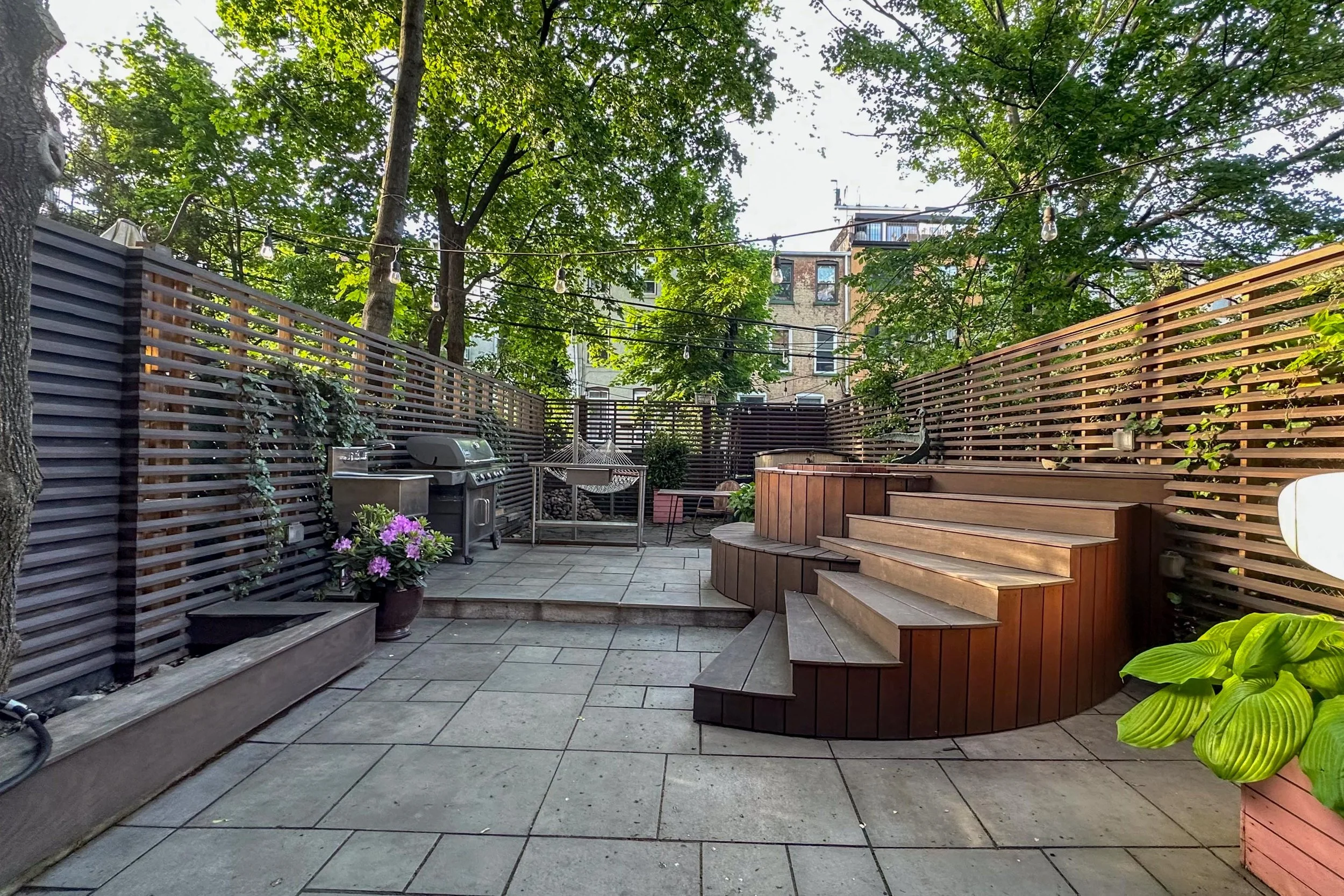 A backyard patio with paved flooring, wooden steps, a grill, hanging string lights, potted plants, and surrounding wooden fencing, with trees and neighboring buildings visible in the background.