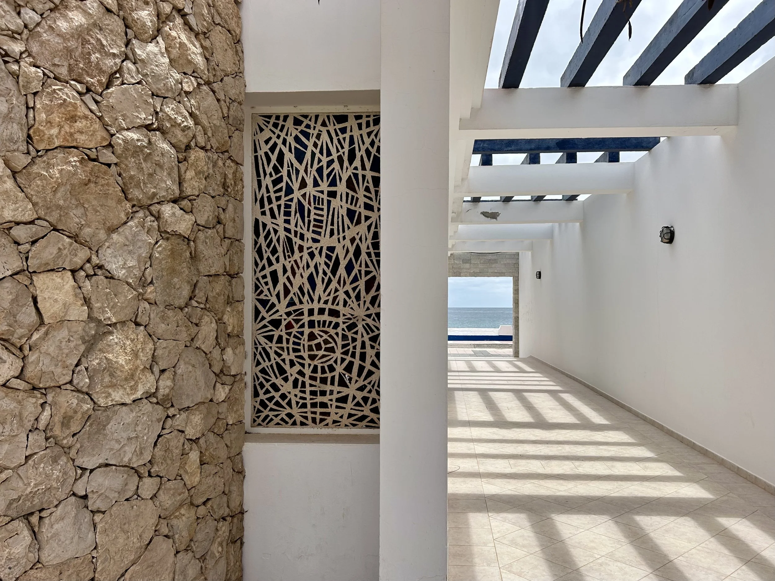 Architectural view featuring a stone wall, a decorative metal screen, white walls, open slats on the ceiling, and a distant view of the ocean.