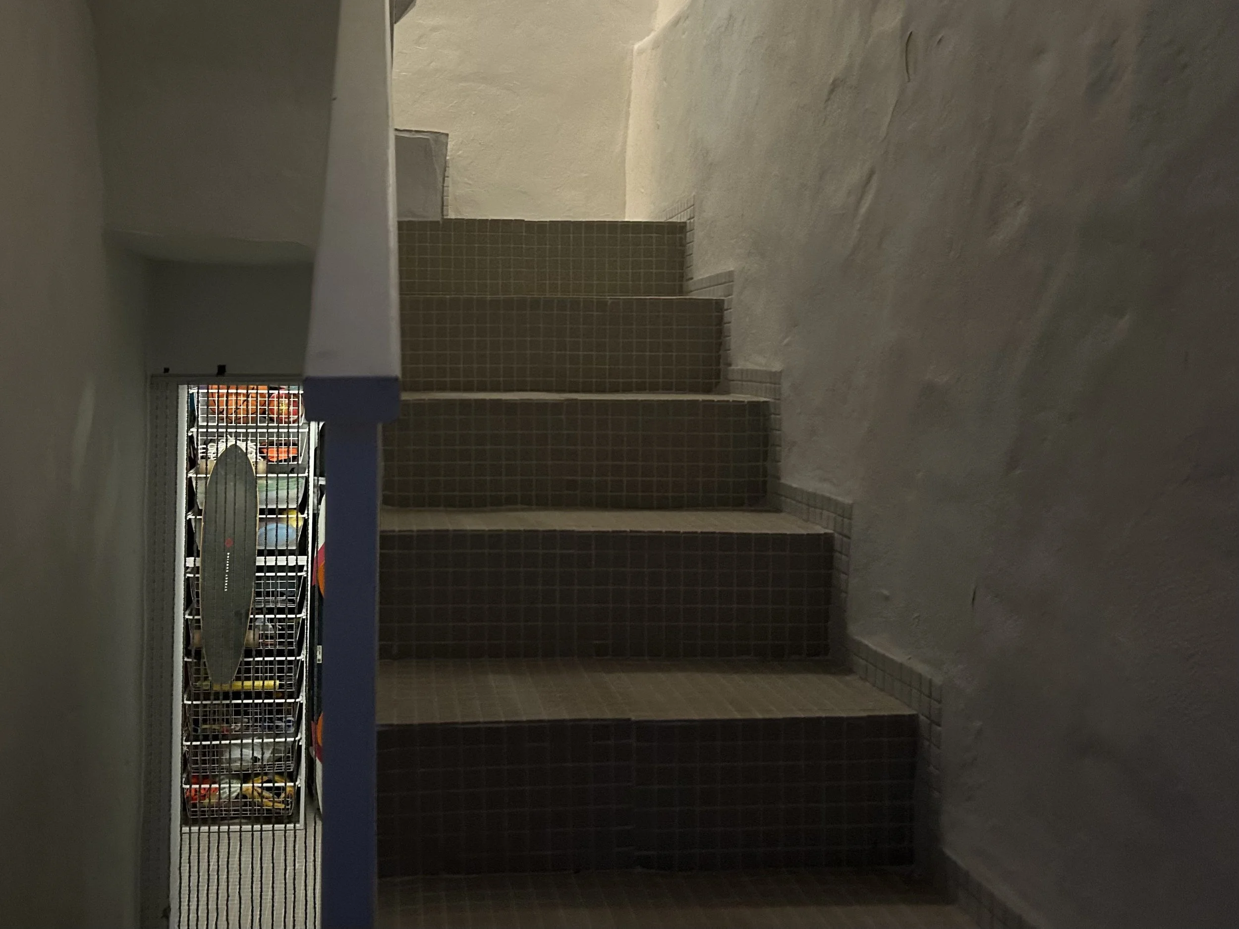 A staircase with brown square tiles on the steps and a laundry basket with clothes on the left side, near a barred window showing a parking lot outside.