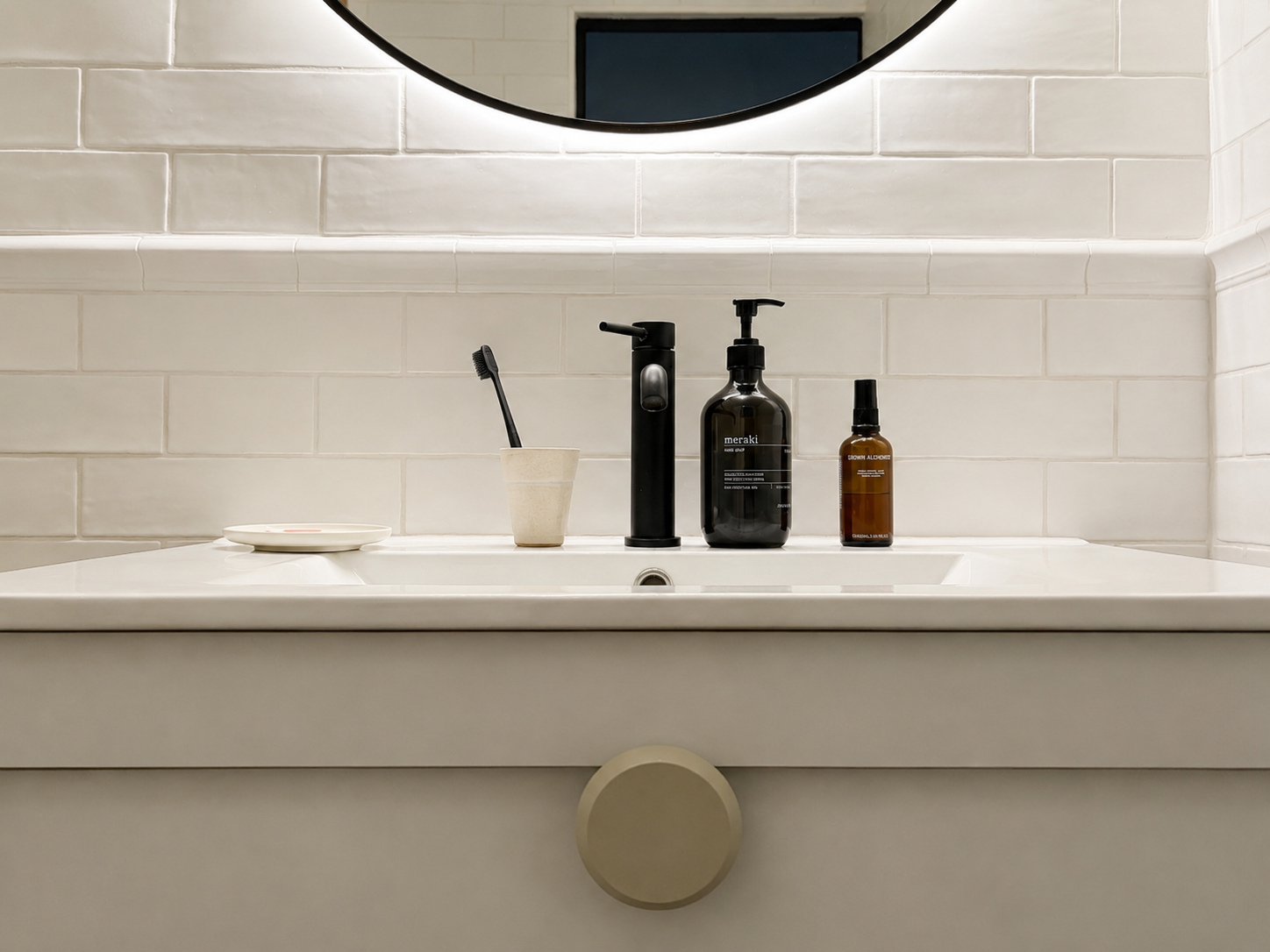 Bathroom sink with a black faucet, decorative soap dispenser, and a dish with a lid, set against a white tiled wall and a round mirror.