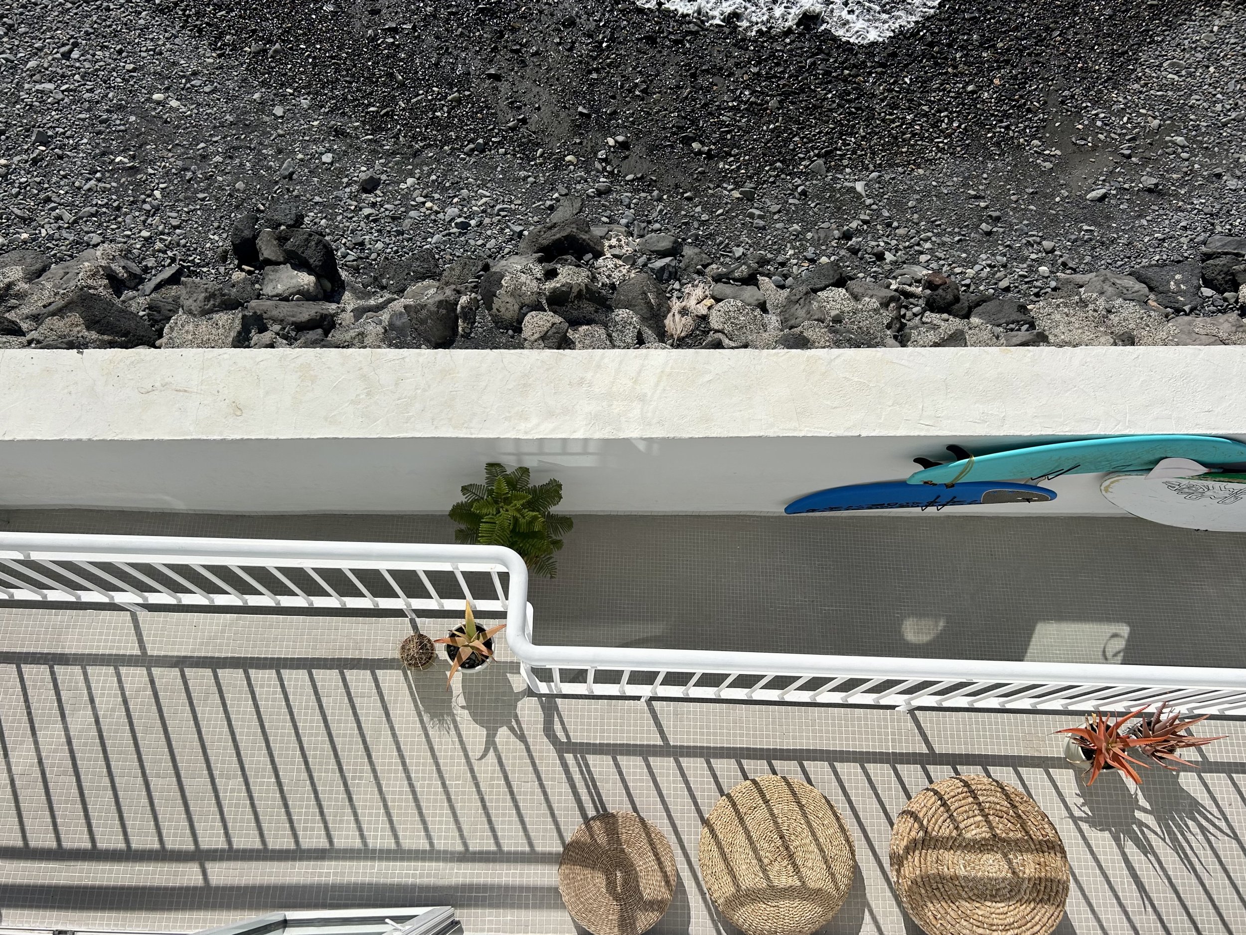 View of a balcony with a white metal railing, a small plant in a pot, two woven hats, and vibrant succulents. Below is a beige tiled floor. A surfboard rests against the railing.