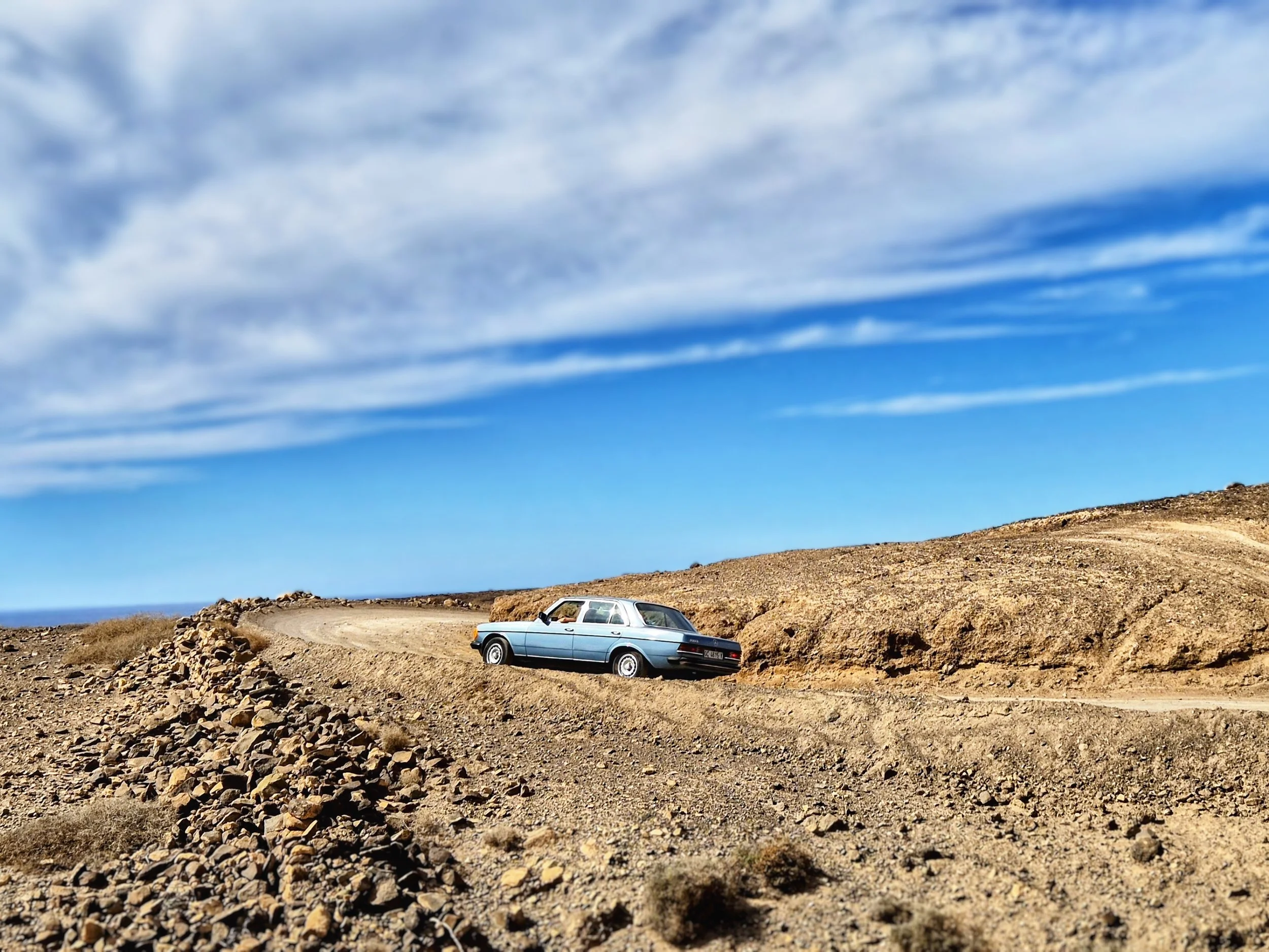 A vintage blue car parked on a dirt road in a rocky, desert landscape with dry grass and no trees, under a bright blue sky with wispy white clouds.