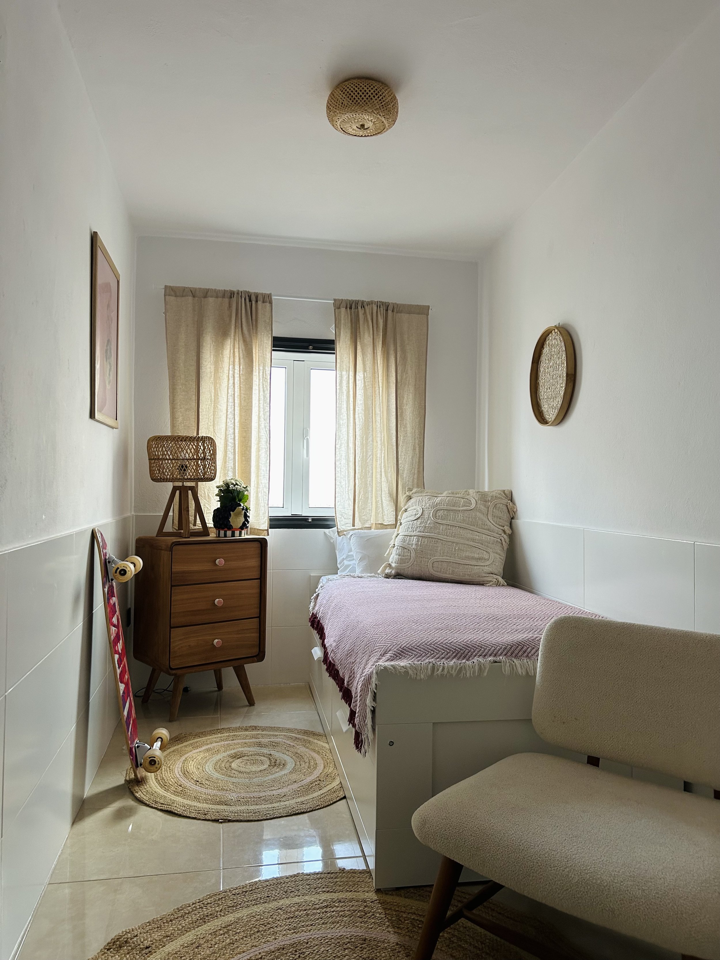 A small bedroom with a twin bed, beige curtains, a window, a wooden dresser with a lampshade and decorative items, a skateboard leaning against the wall, two oval wall decorations, a beige chair, and a round woven rug on a tiled floor.