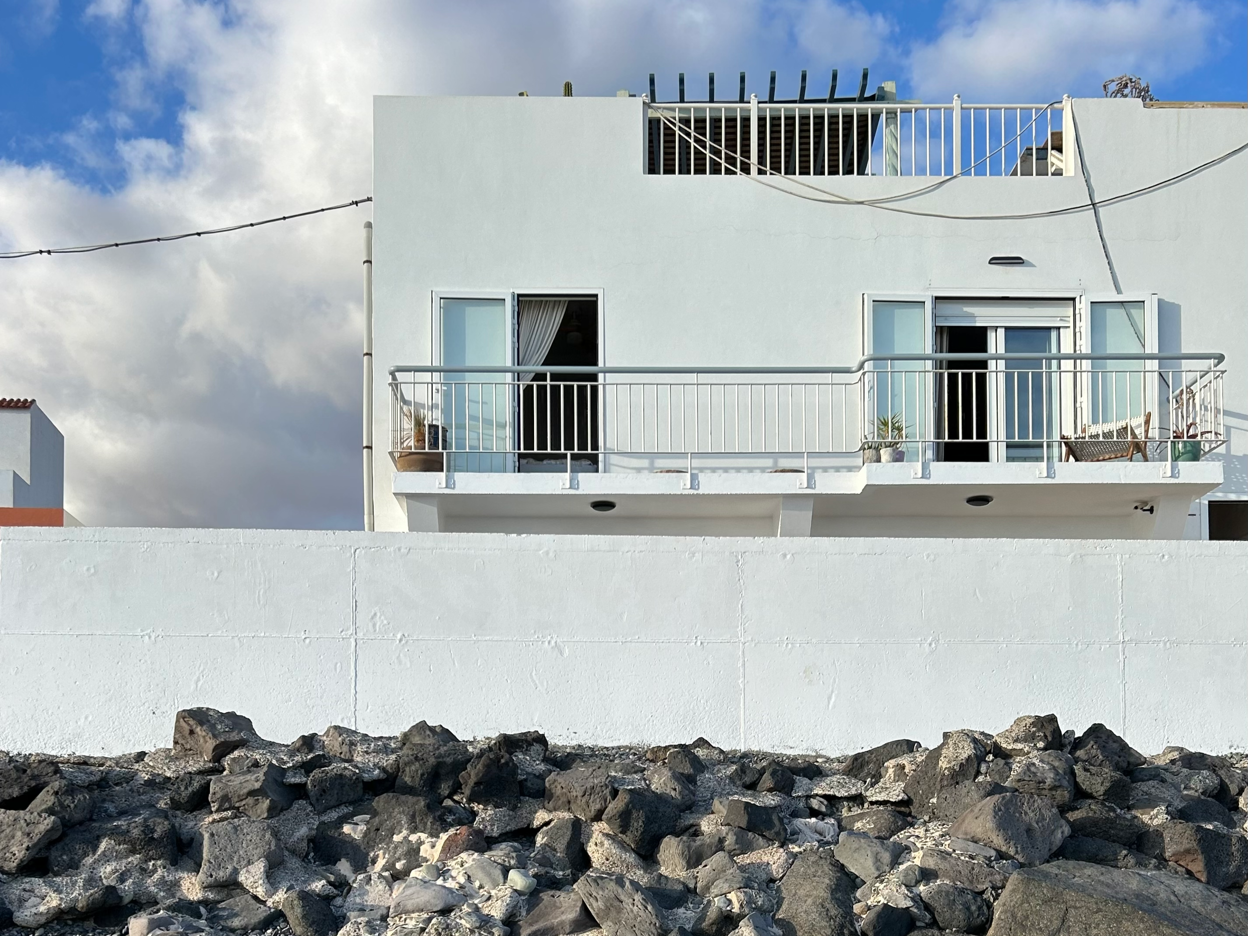 A white, modern building with balconies and large windows, set against a partly cloudy sky, with rocks and volcanic stones in the foreground.
