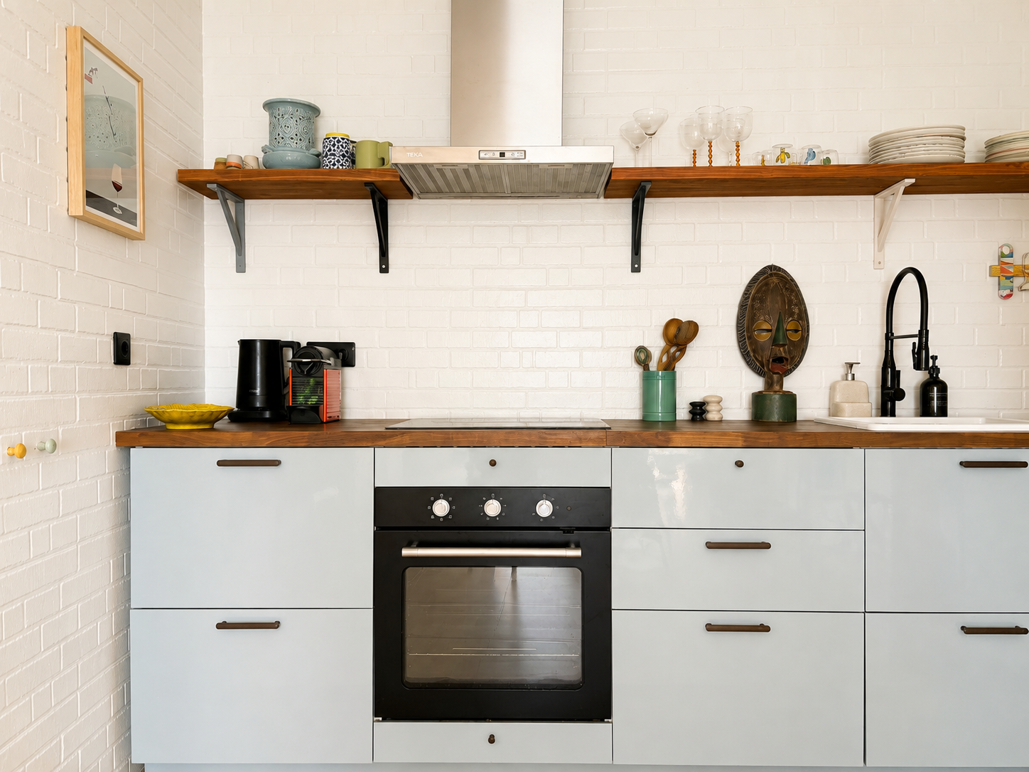 A kitchen with a white brick wall, wooden and metal floating shelves holding cups, glasses, plates, and bowls. There is a black stove with a white oven beneath and various kitchen utensils and containers on the countertop.