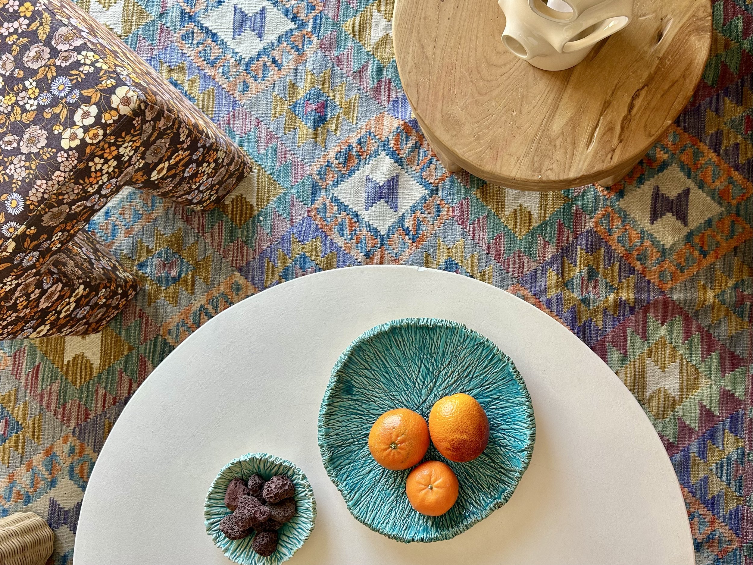 A white round table with four colorful decorative bowls, three orange fruits in one bowl, a few dark purple snacks in another, and a teal-colored bowl with a black sketch of tree branches. The table is on a multicolored patterned rug, and part of an armrest and white chair are visible in the image.
