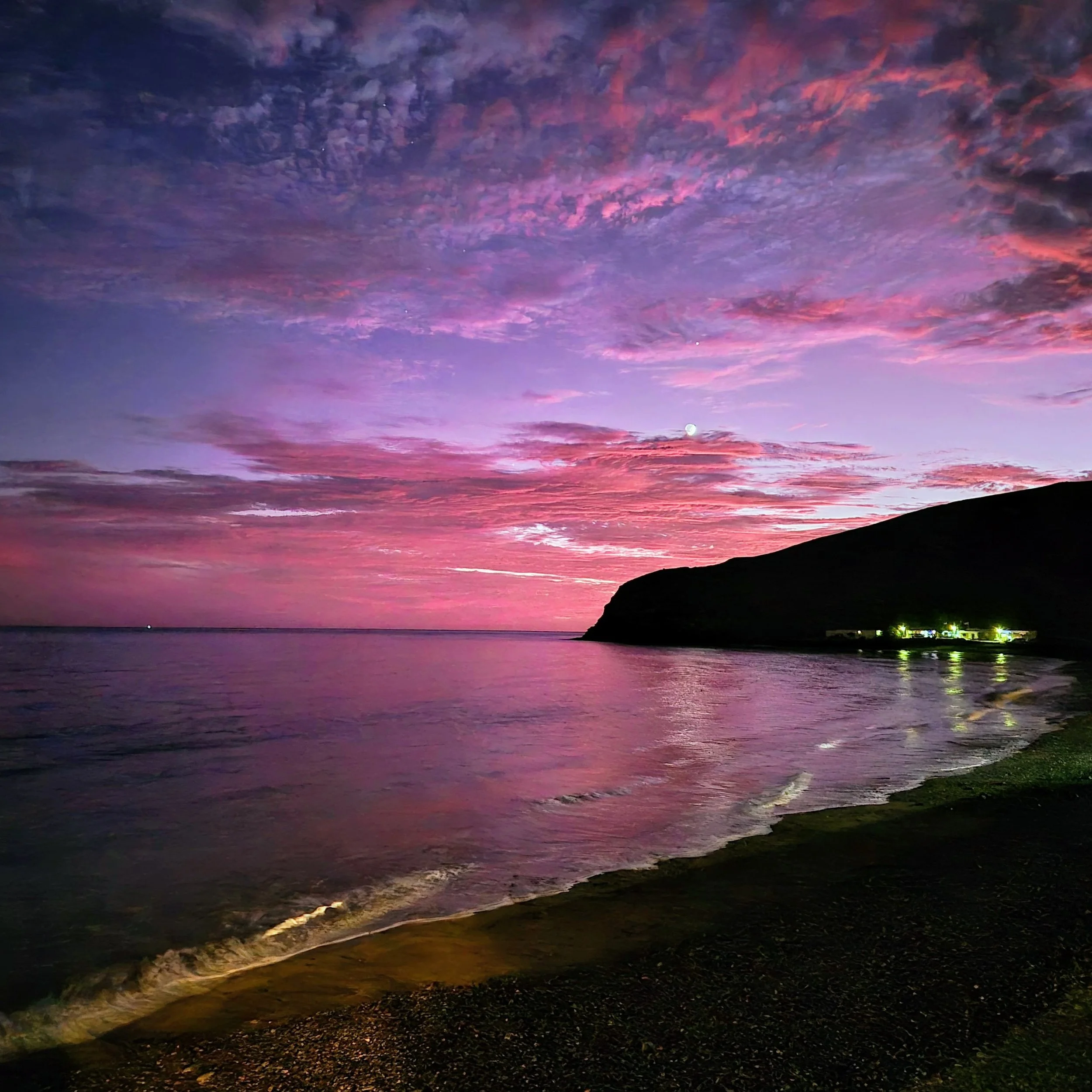 Colorful sunset sky over a calm ocean with a silhouette of a mountain or island in the distance and a few lights near the shoreline.