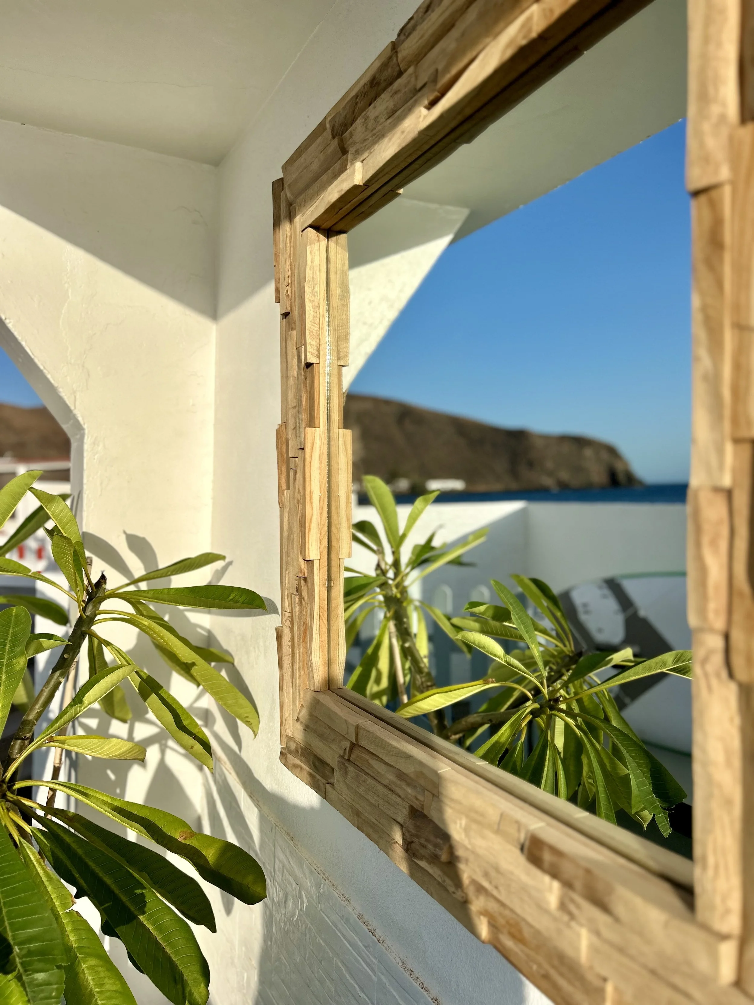 Wooden window frame partially opened on a white wall, with green plants outside and a mountain in the background under a clear blue sky.