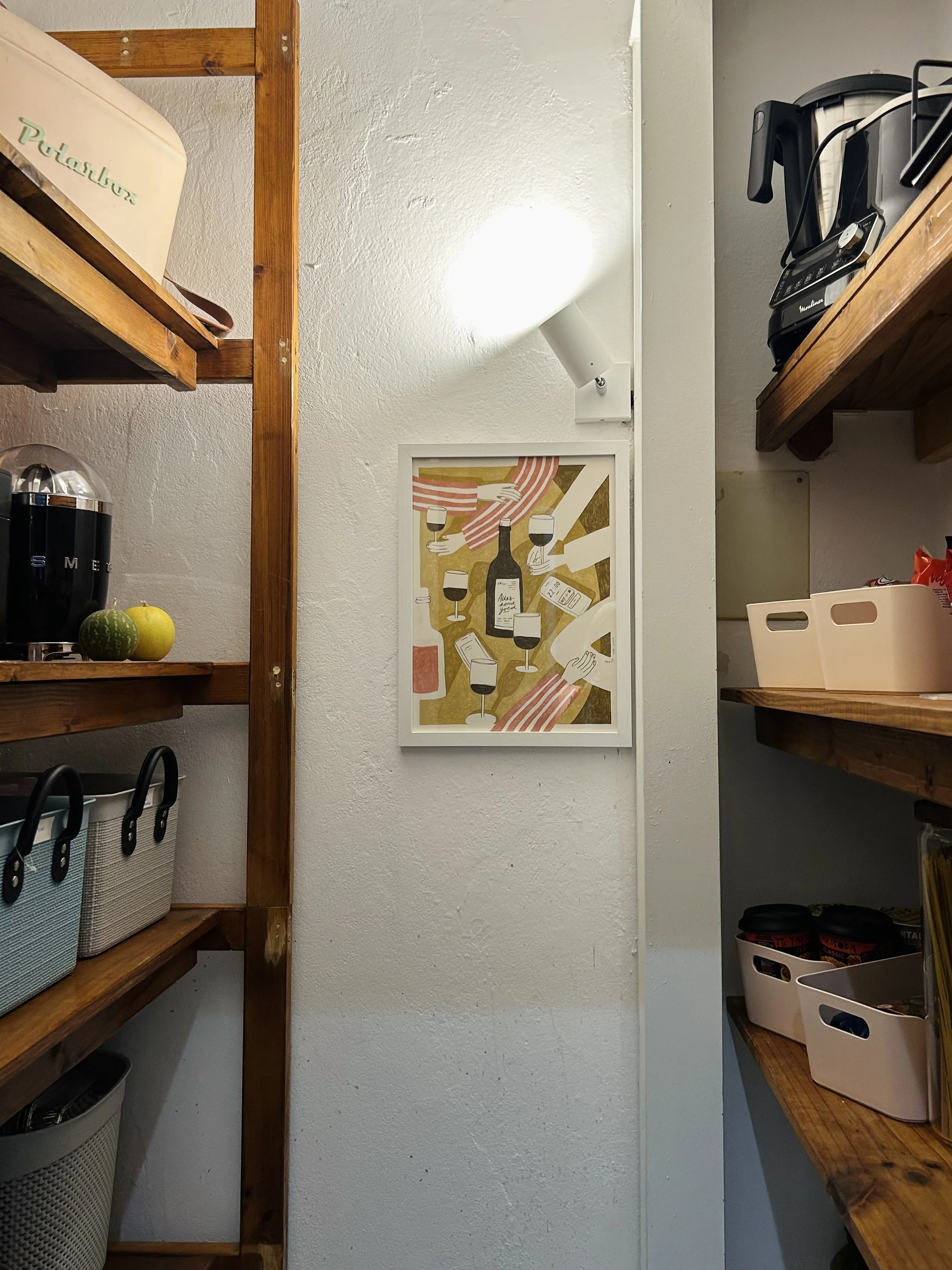 A corner of a kitchen with wooden shelves and a white wall. On the upper left shelf, a beige container with green writing. On the right shelf, a black and silver coffee maker. Framed art on the wall depicts wine glasses, wine bottles, and hands reaching for the glasses.