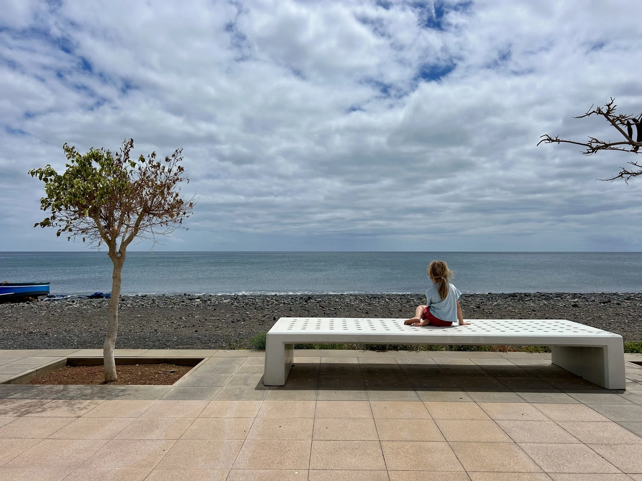 A young girl with blonde hair sitting on a white bench facing the ocean, with trees and cloudy sky in the background.