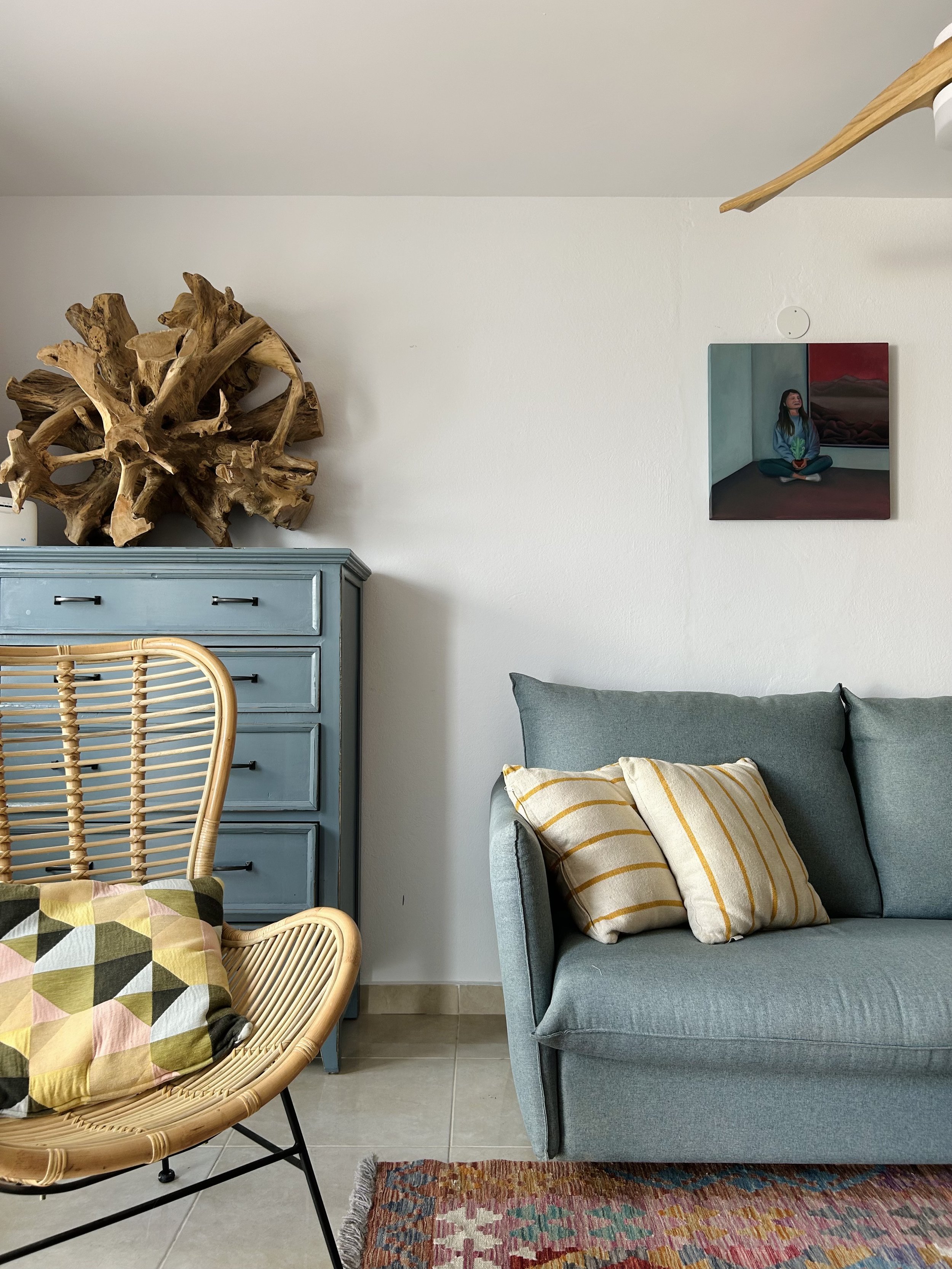 Interior living room with a light blue cabinet, a wicker chair with a multicolored cushion, a gray sofa with a yellow striped pillow, a colorful rug, and wall art depicting a woman sitting in a room with mountains outside.