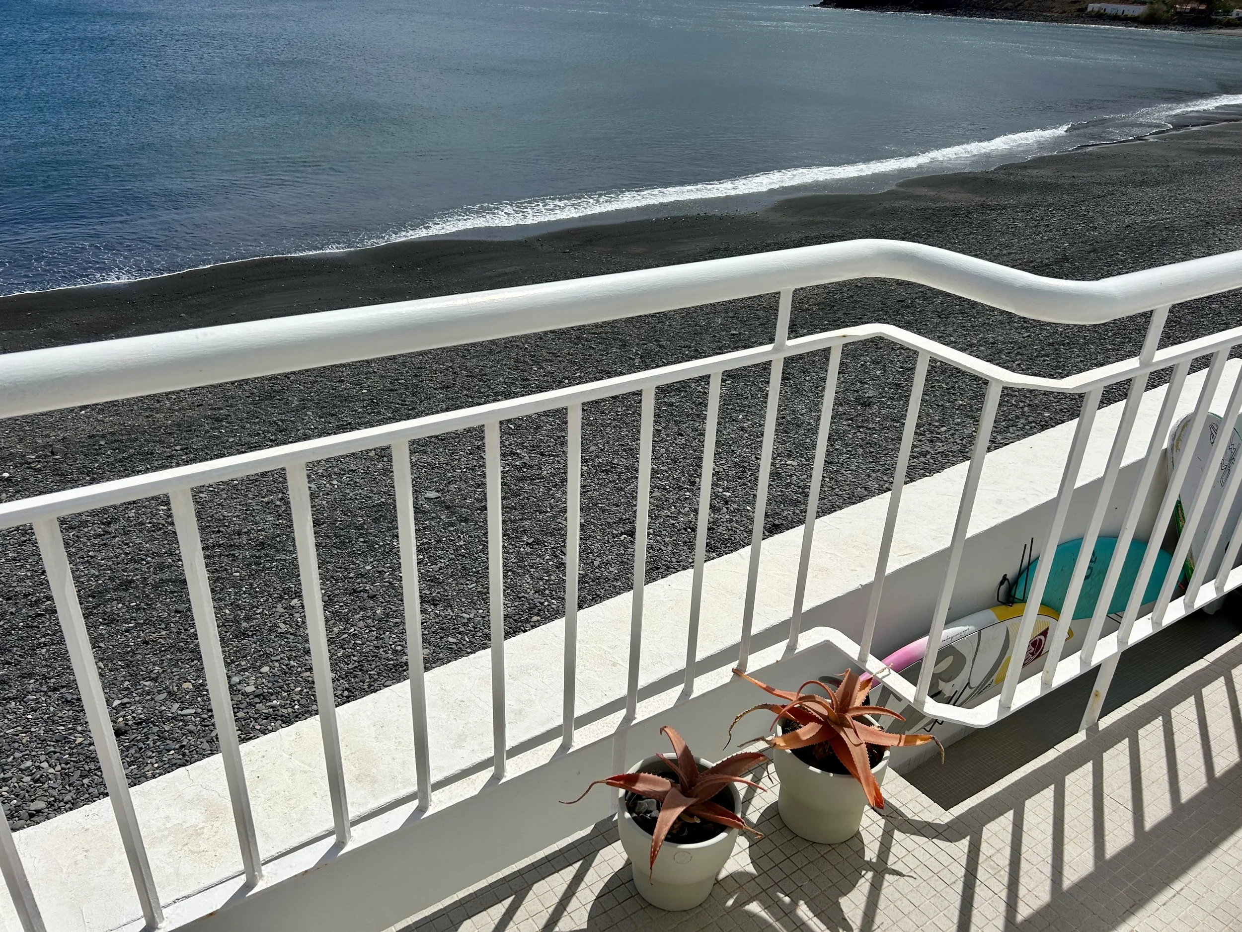 View of a beach with dark sand, gentle waves, and a railing on a balcony or porch. There are potted plants and a skateboard near the railing.