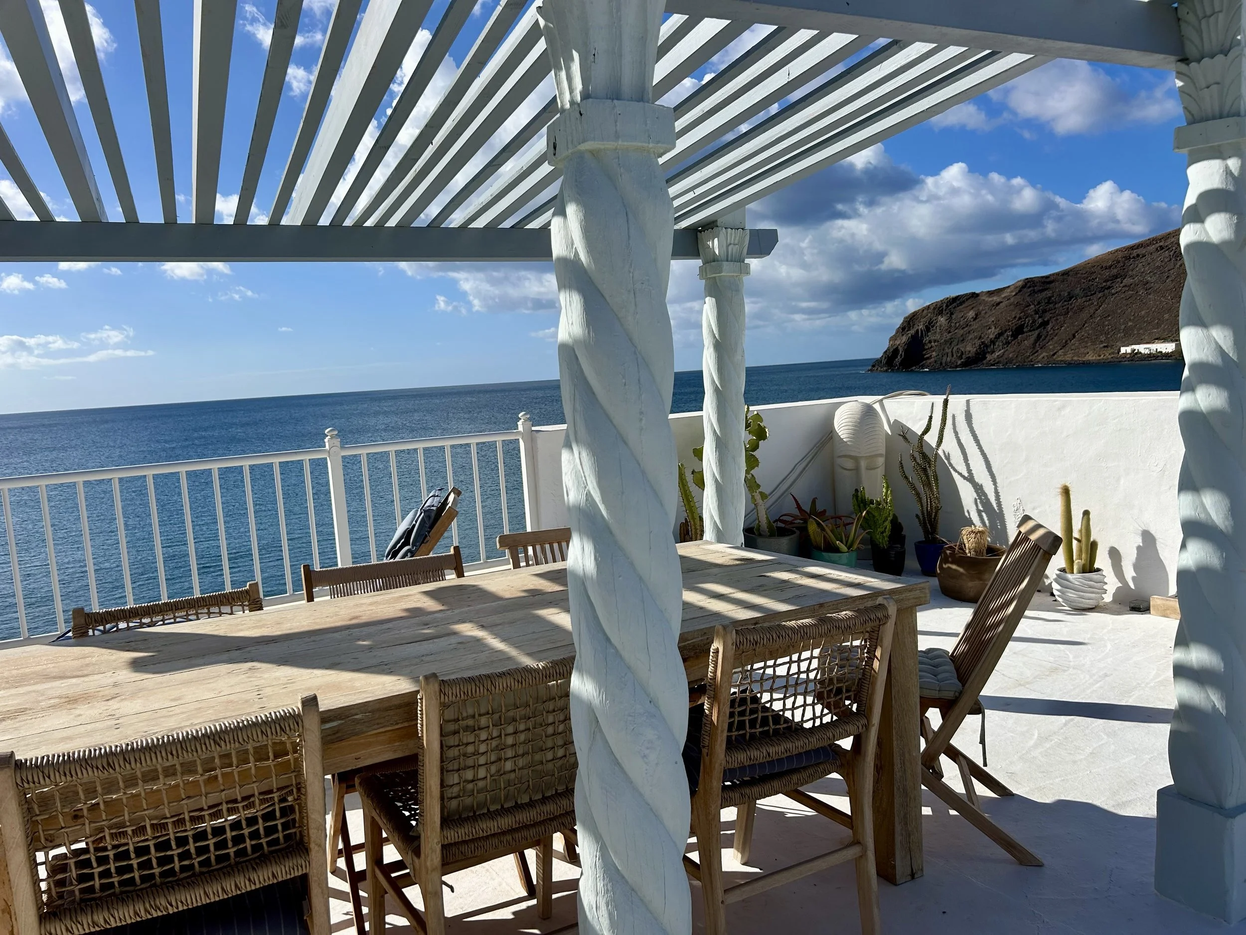A seaside outdoor dining area with a wooden table and woven chairs, surrounded by white walls and railings, overlooking the ocean with a rocky hillside in the background under a partly cloudy sky.