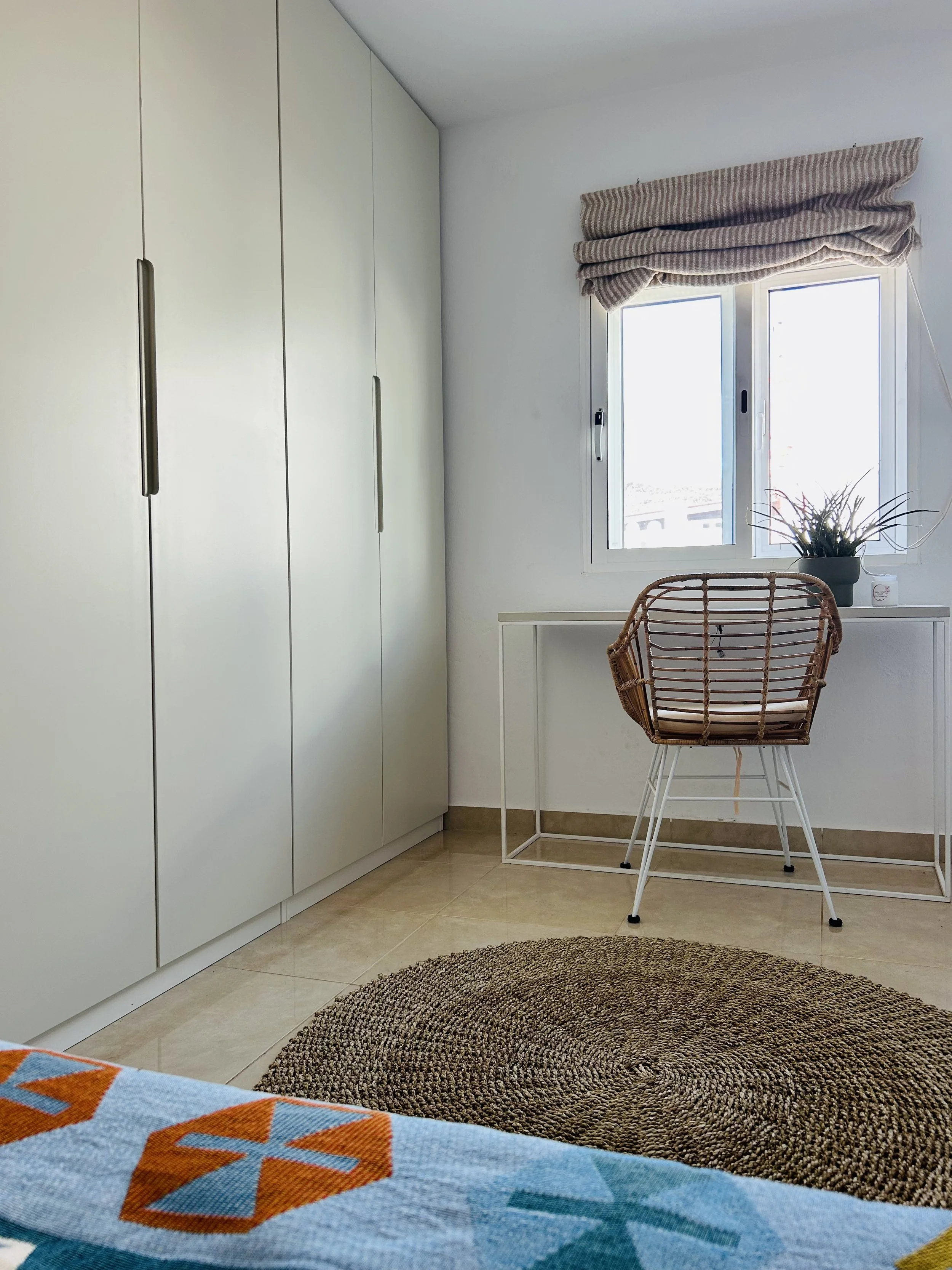 A corner of a room with a white wardrobe on the left, a window with a striped beige and brown curtain, a rattan chair with white metal legs in front of the window, a small white desk beneath the window, and a potted plant on the desk. The floor is covered with beige tiles and a round brown woven rug.