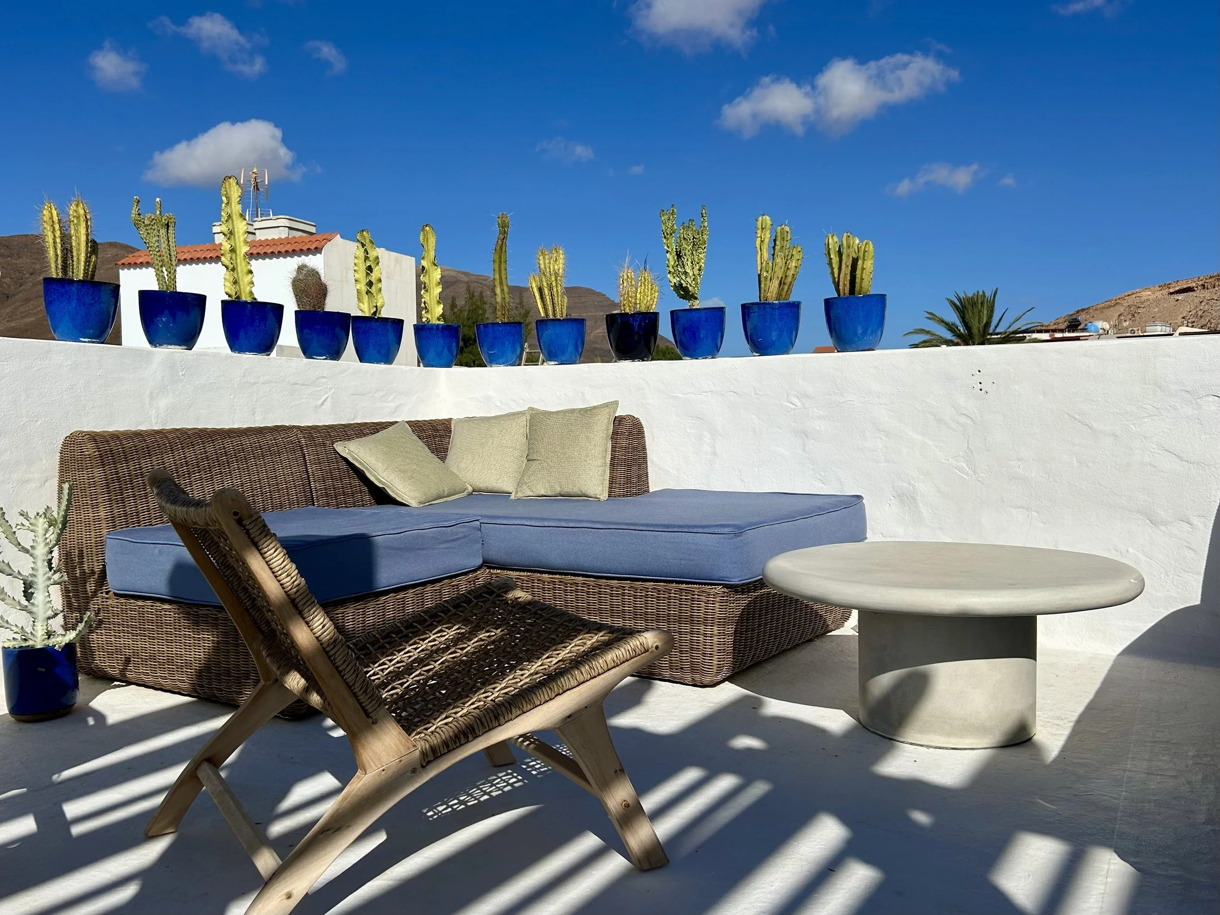 Outdoor patio with blue cushions, woven chair, white round table, potted cactus plants in blue pots, white wall, background mountains, clear blue sky with a few clouds.