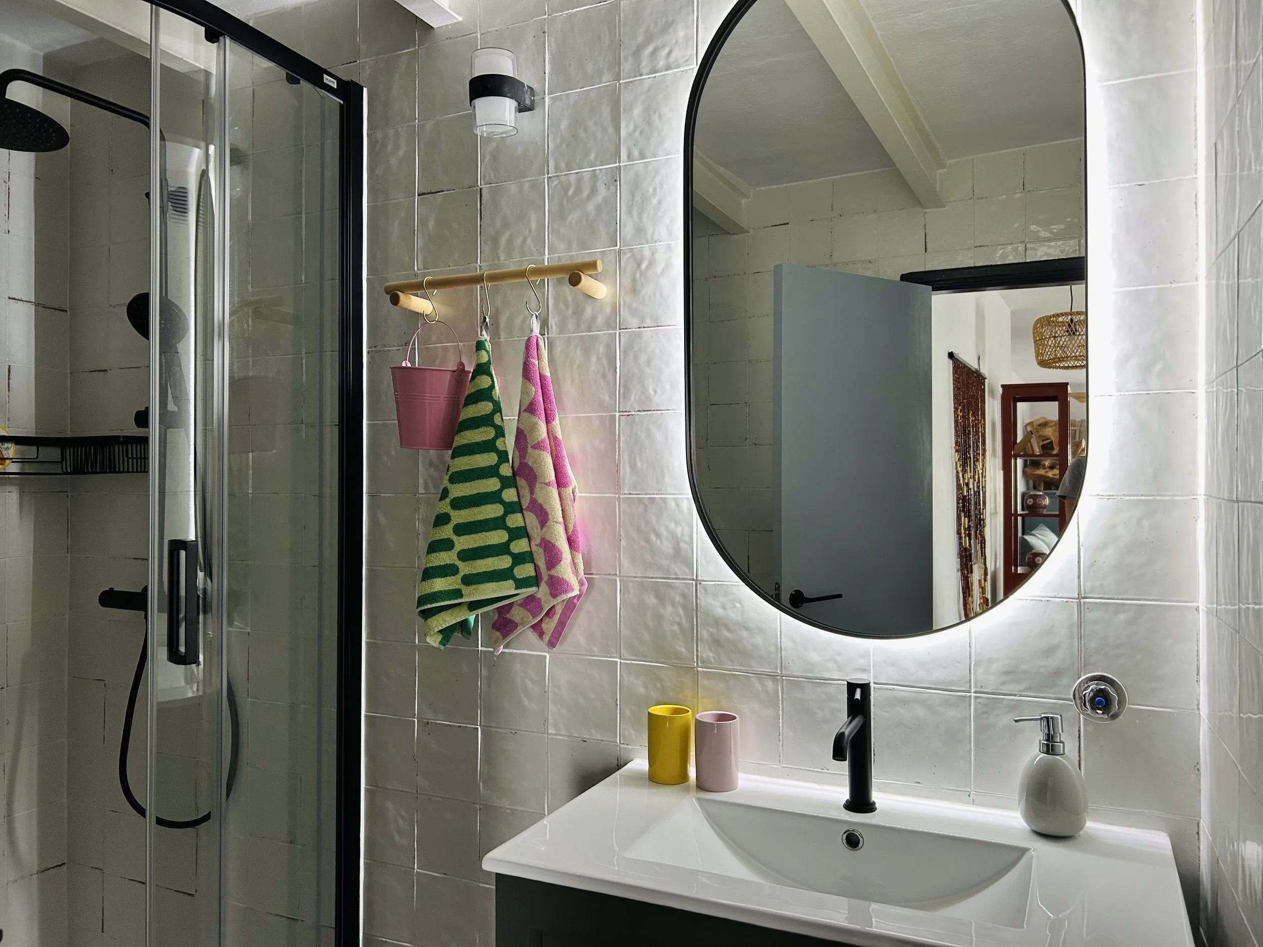 A bathroom with a large round mirror above a white sink, black faucet, and beige tiled walls. Two colorful towels hang on a wooden rack, and three vases are on the sink counter. A soap dispenser and exposed shower area are visible.