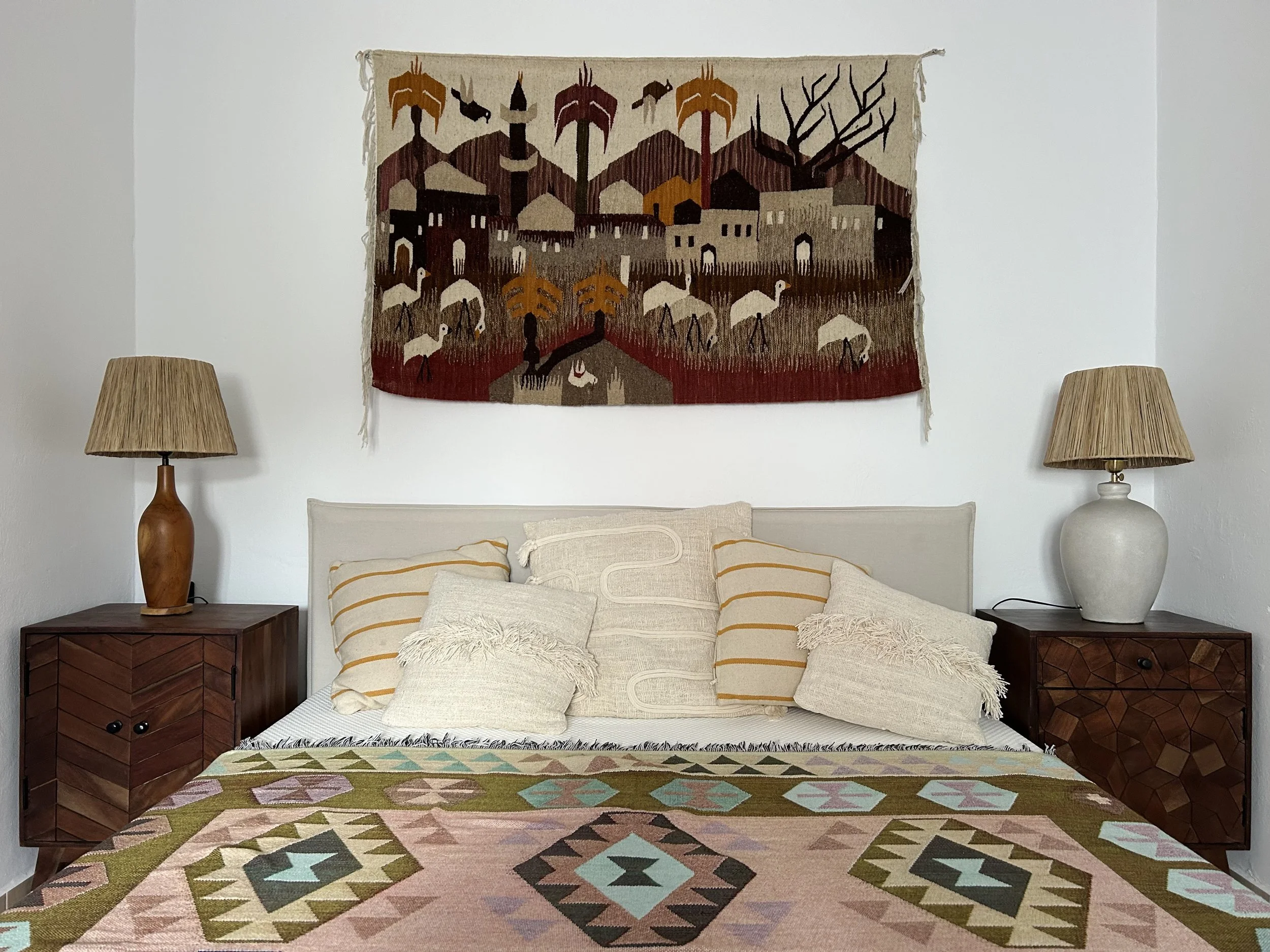 Living room with a large Southwest-style geometric patterned rug on the floor, cream-colored couch with pillows, and wooden nightstands with lamps on each side; a colorful woven wall hanging depicts a desert scene with mountains, trees, and animals.