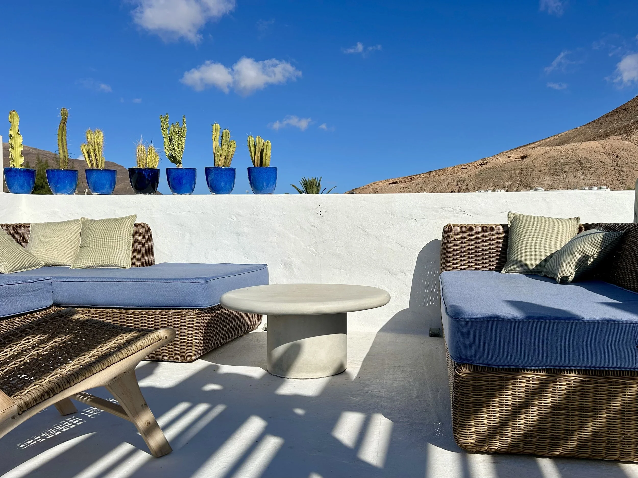 Rooftop terrace with wicker sofas, a small round table, blue planters with cacti, and a view of arid mountains under a blue sky with few clouds.