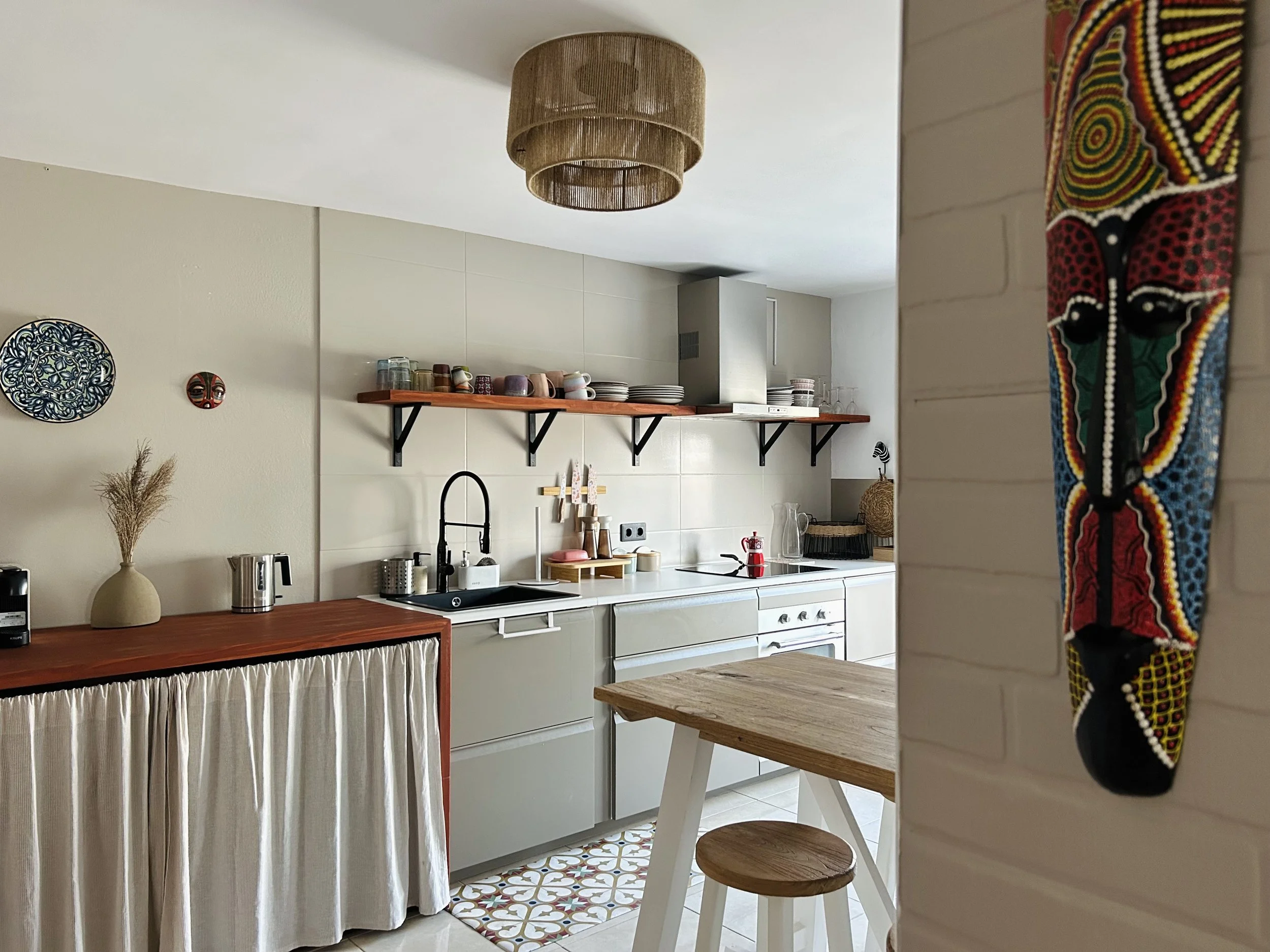 A cozy kitchen with a wooden table and a wooden bar stool. The kitchen has hanging shelves with dishes and glassware, a modern sink with a black faucet, and decorative wall art. A woven ceiling light and a patterned floor tile are also visible.