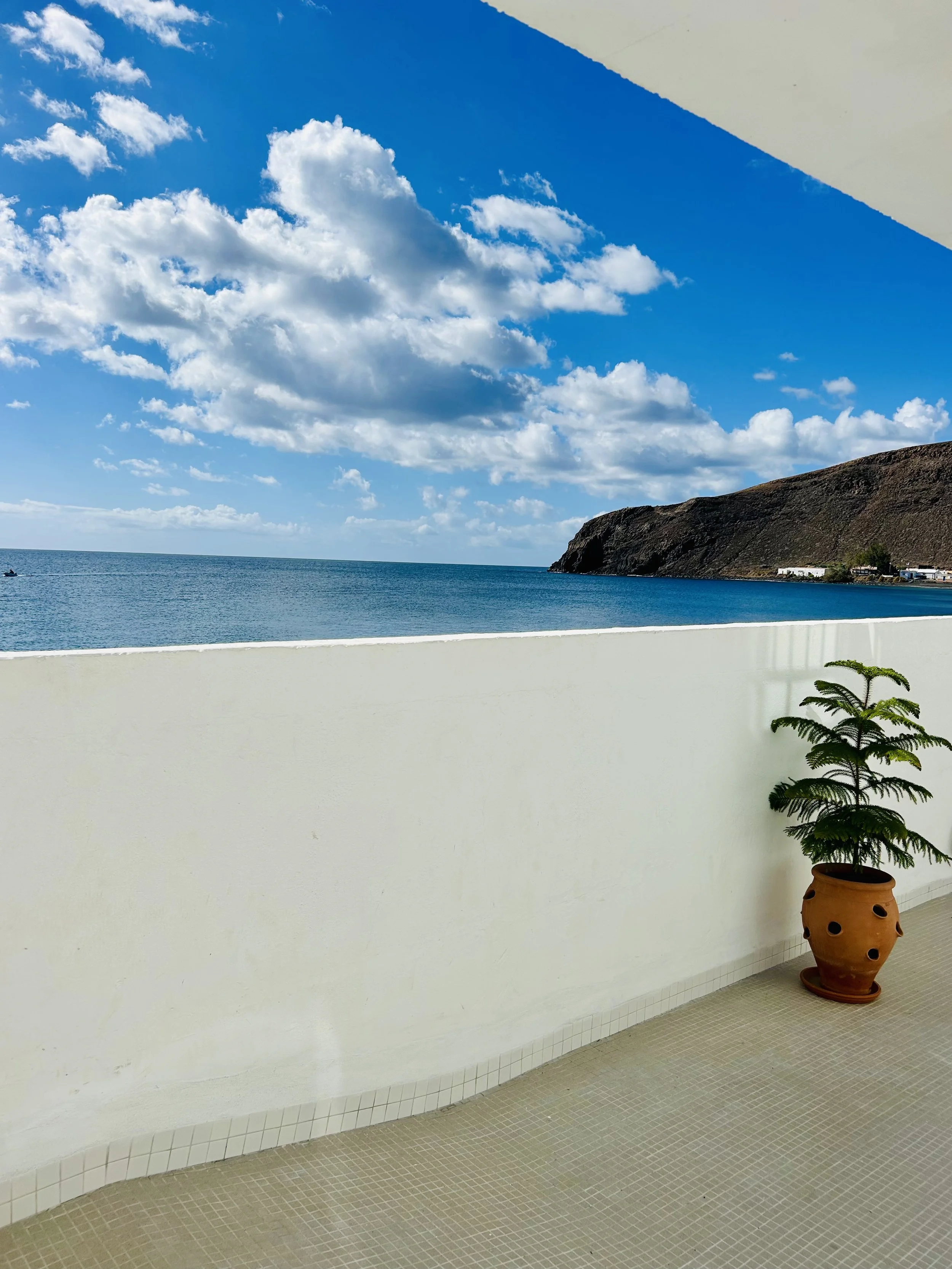 A view from a balcony or terrace showing the ocean with a rocky hill and buildings in the distance. The sky is partly cloudy with white clouds and blue sky, and a potted plant is placed on the balcony with a white wall and tiled floor.