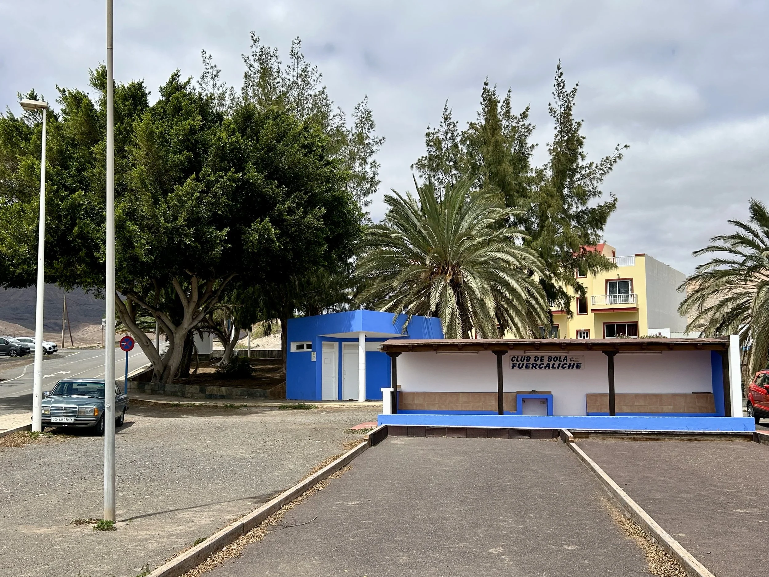 An empty parking lot with a small blue and white structure labeled "Club de Bola Fuercaliche" in front of a mix of tall trees, a yellow building, and street lamps under a cloudy sky.
