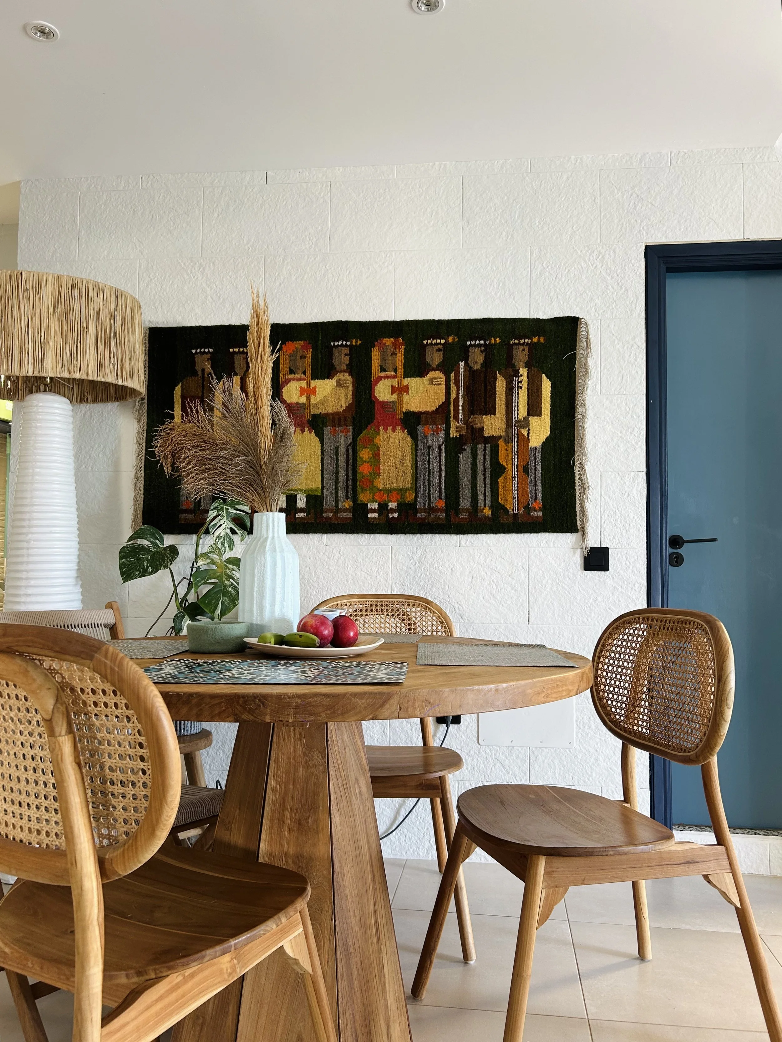 A round wooden dining table with five wooden chairs around it, one with a cane backrest. On the table, there is a white plate with three apples and two bananas, two placemats, and a gray pot with dried floral arrangements. To the left, there is a tall white vase with a dried plant. The background has a wall with a woven tapestry depicting four stylized people in colorful clothing, and a blue door on the right.