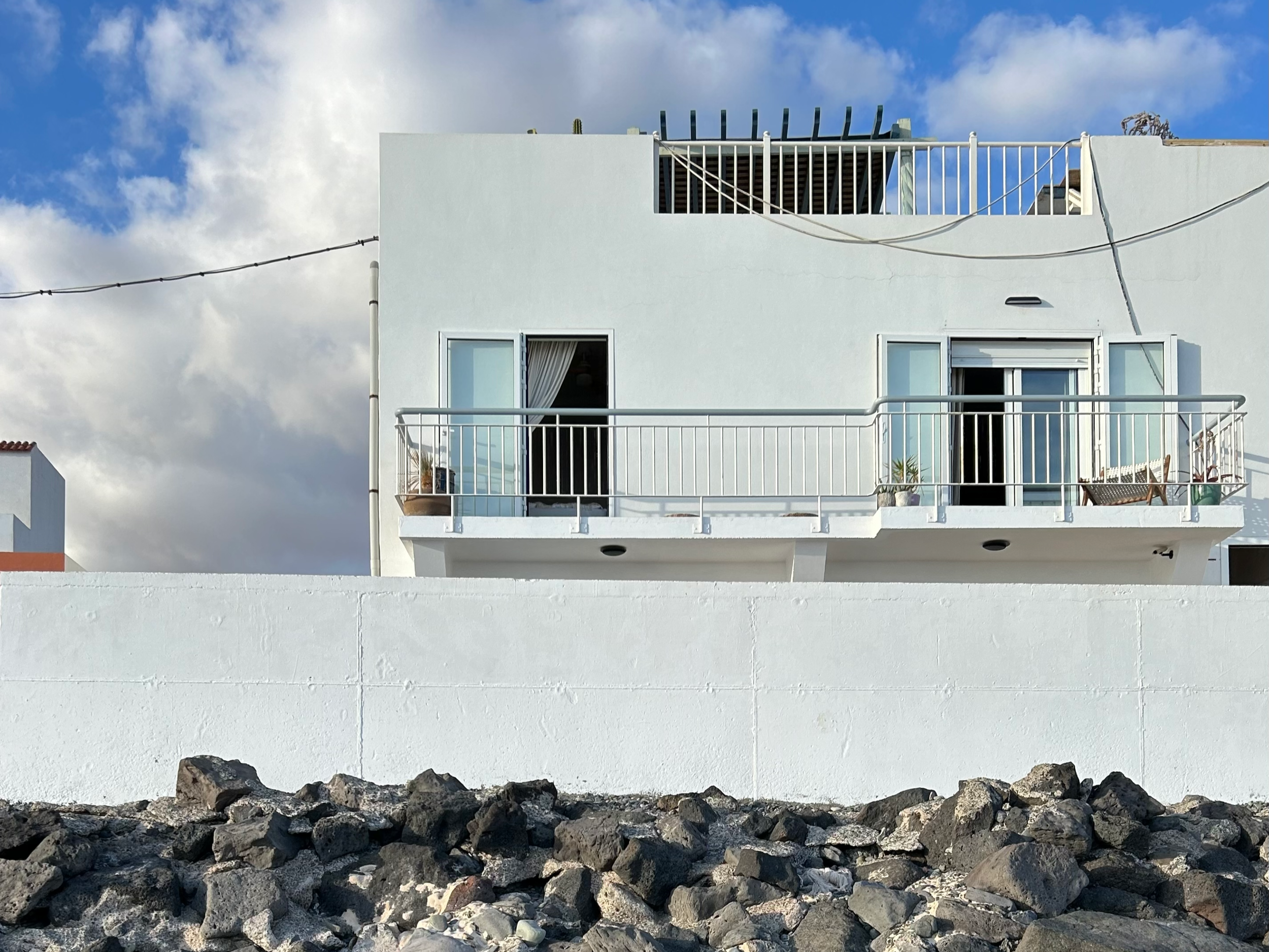 White modern building with balcony, sliding glass door, and rooftop structure, against blue sky with clouds, built on rocky terrain.