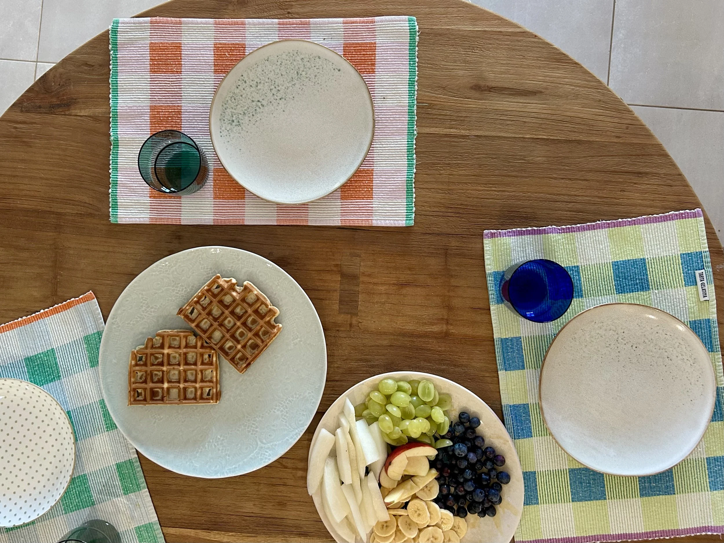 A round wooden table with a checkered placemat, holding plates of waffles, fresh fruit, and bowls of grapes, bananas, and berries. There are colorful cloth napkins underneath some plates and glasses of water.