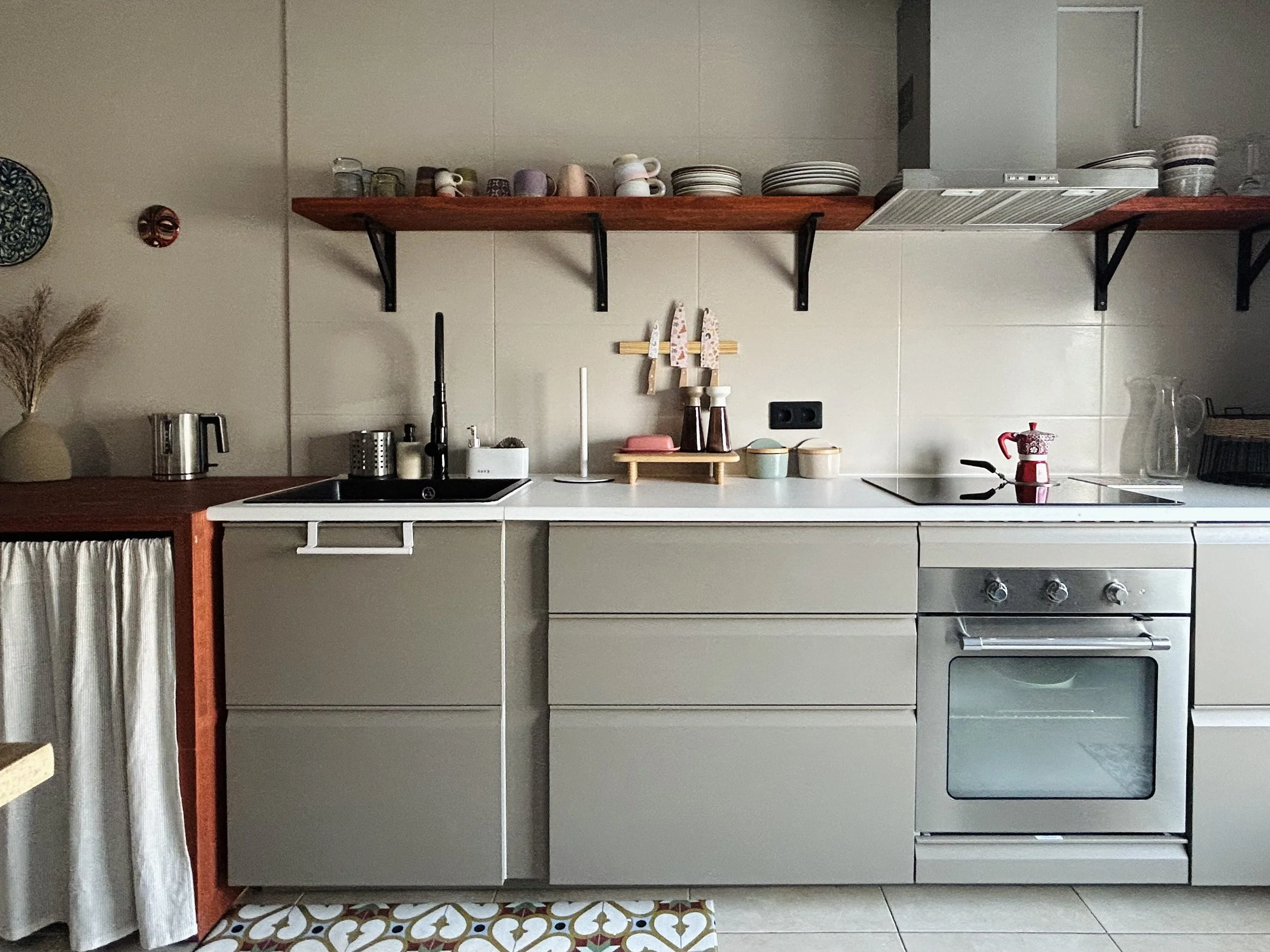 Kitchen with beige cabinets, open shelves with cups and plates, black sink, and stovetop with oven, decorated with patterned floor tiles.