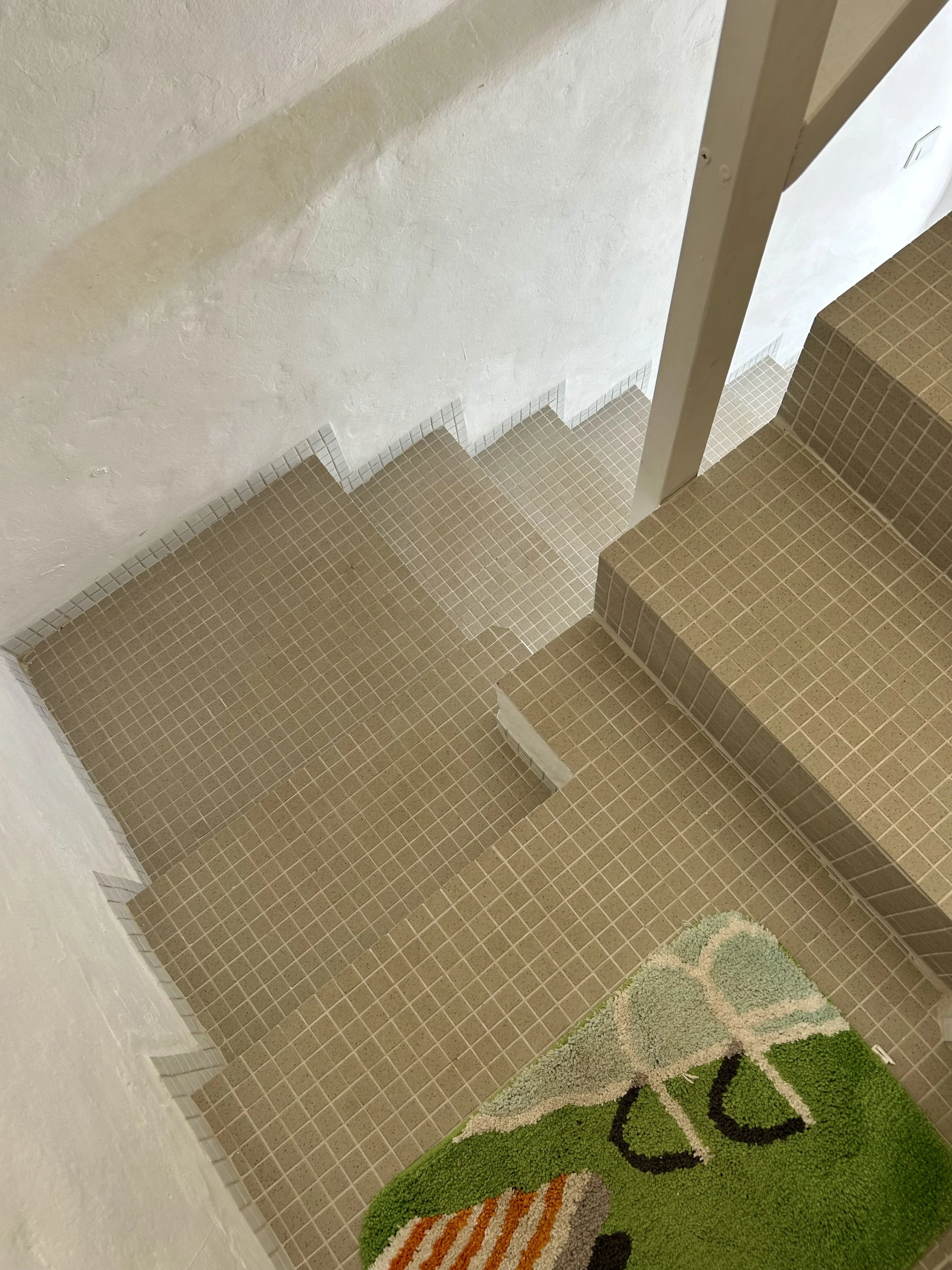 View of a tiled staircase with small beige square tiles, a white wall with a textured finish, and a partial view of a colorful rug with a floral design on a green background.