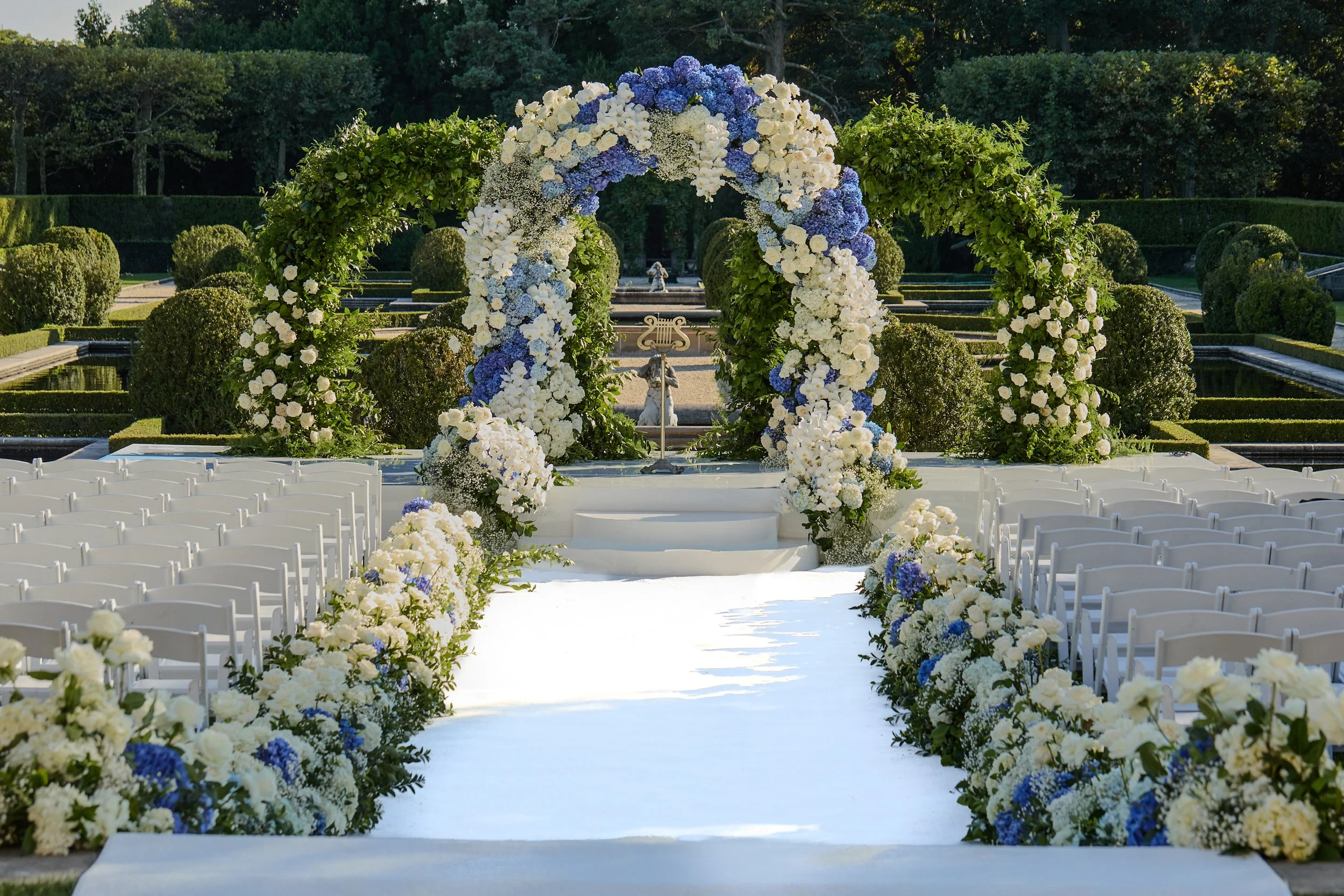 Set within the gardens of Oheka Castle, the ceremony was a study in elegance and restraint. A lush aisle of white florals guided guests toward a striking floral arch composed of blue and ivory blooms, designed to feel both organic and architectural. 