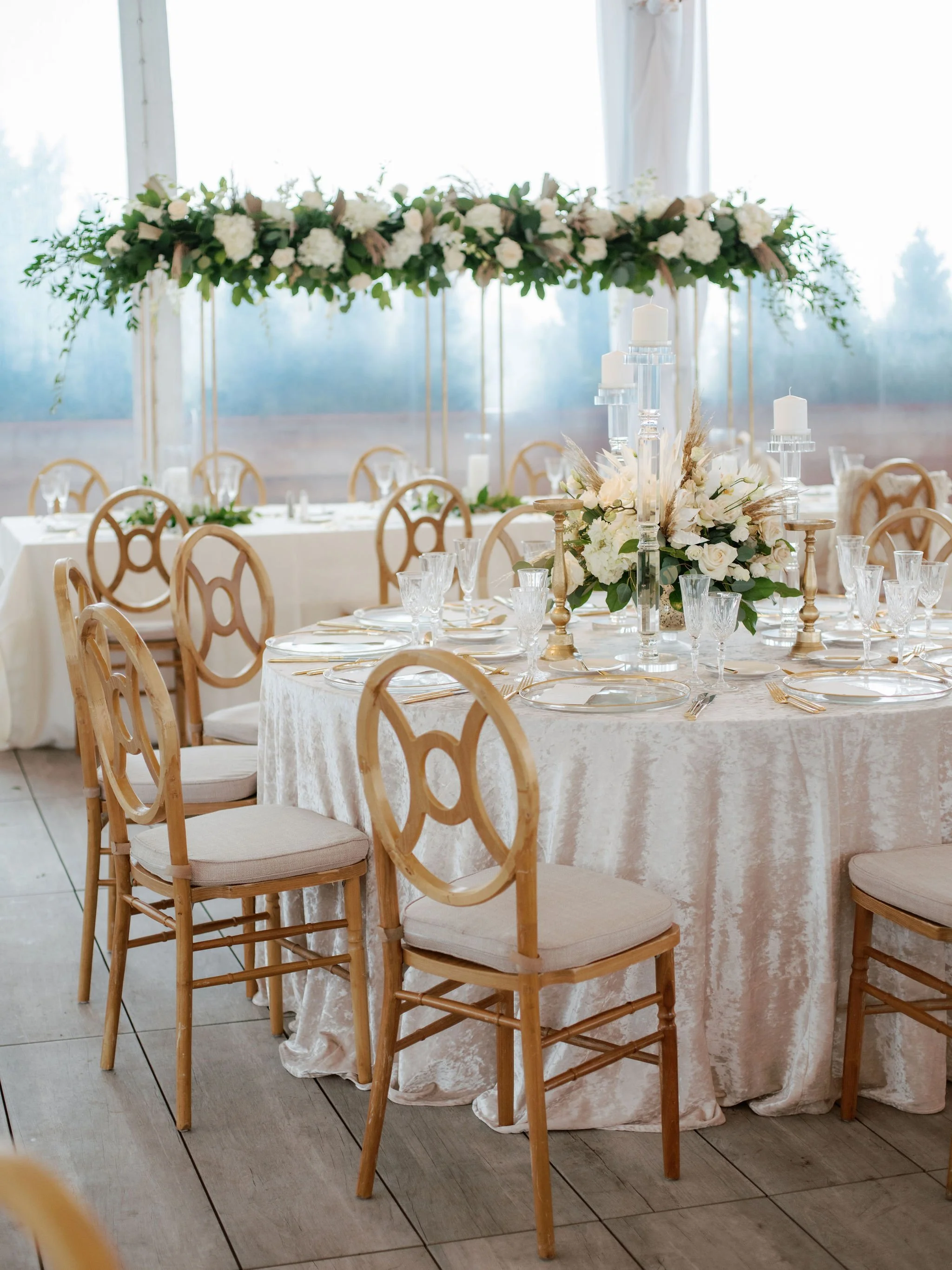 Elegant table decorated with white flowers, candles, and glassware for a wedding reception or event, with a high floral centerpiece and light wood chairs around the table.